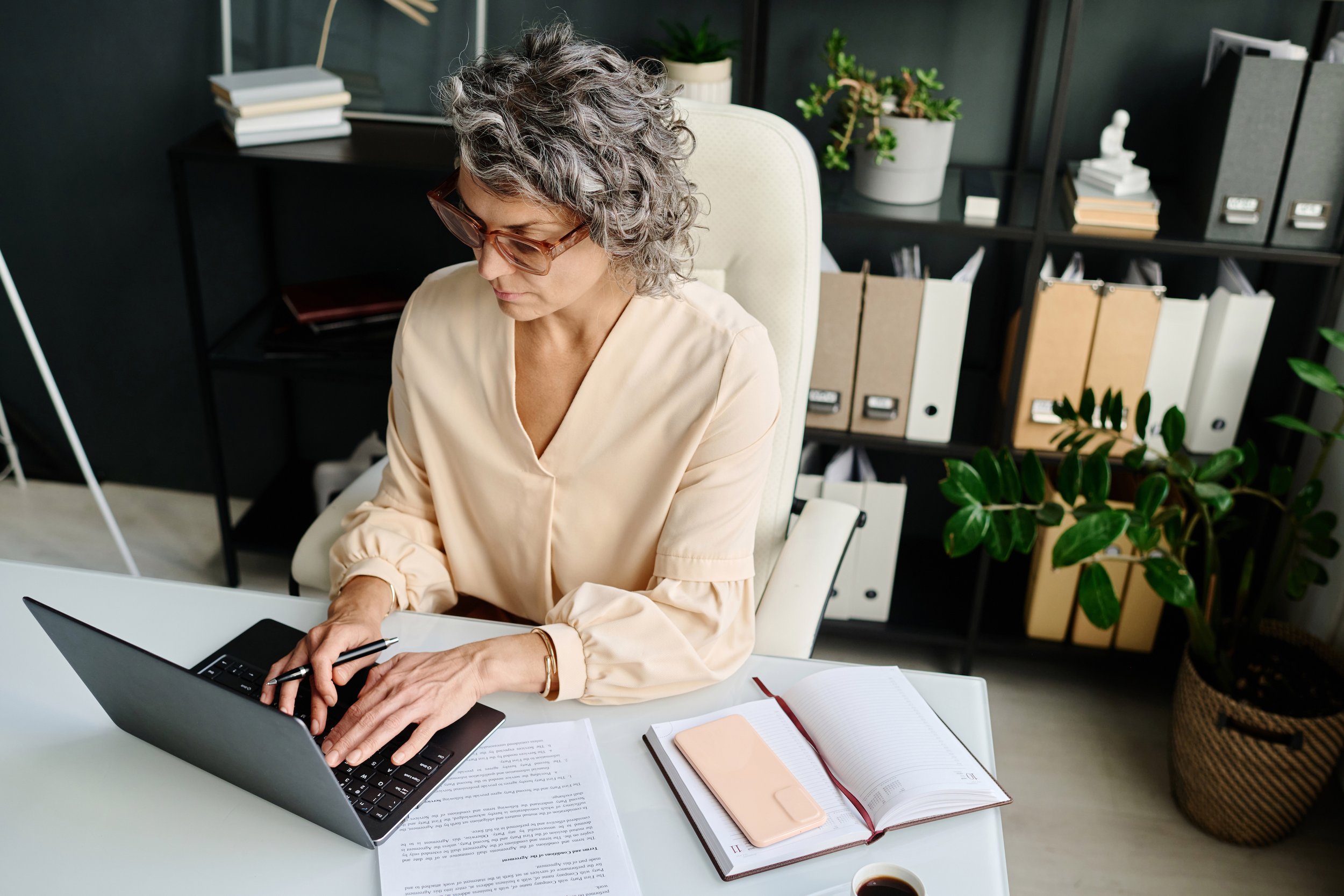 An older woman with curly gray hair and glasses working on a laptop at a white desk in an office. The desk has an open notebook, a closed notebook, a cup of coffee, and papers. Behind her are shelves with books, binders, and plants.