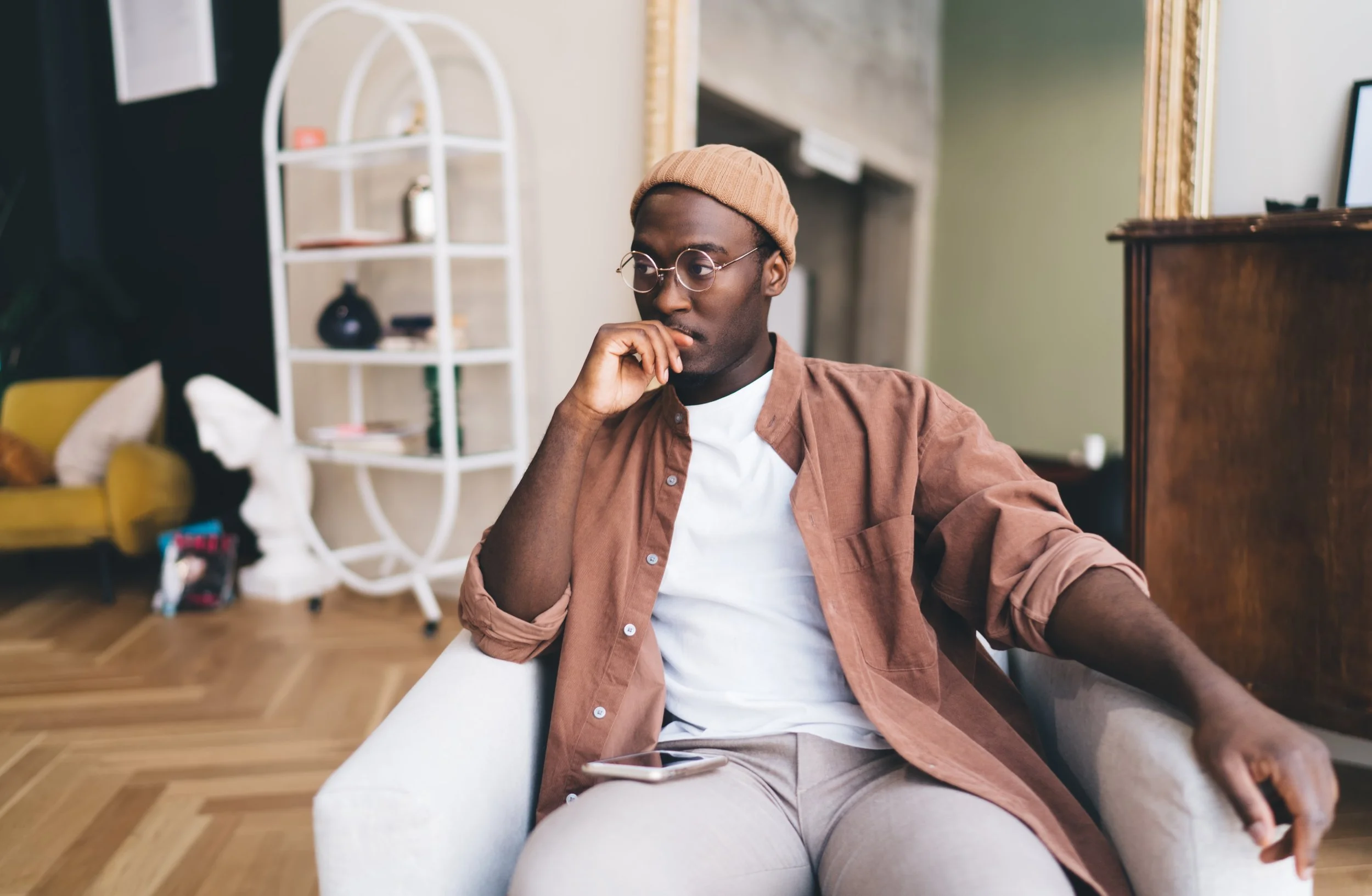 A young man with dark skin, round glasses, and a beige beanie, is sitting on a white couch, resting his chin on his hand, in a cozy home interior. He is wearing a brown shirt over a white t-shirt and beige pants. There is a smartphone on his lap. The background features a white bookshelf with decorative items, a yellow armchair, and a wooden dresser.