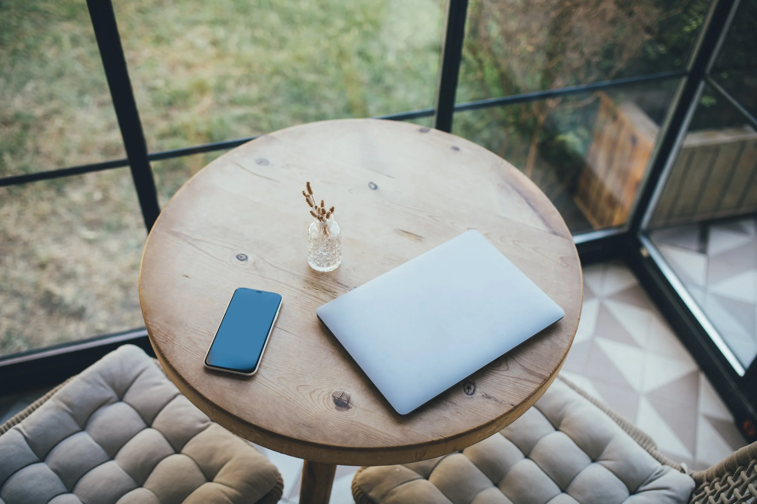 A round wooden table with a smartphone, closed laptop, small vase with dried flowers, and a piece of paper, situated in front of a large glass window showing a grassy outdoor scene.
