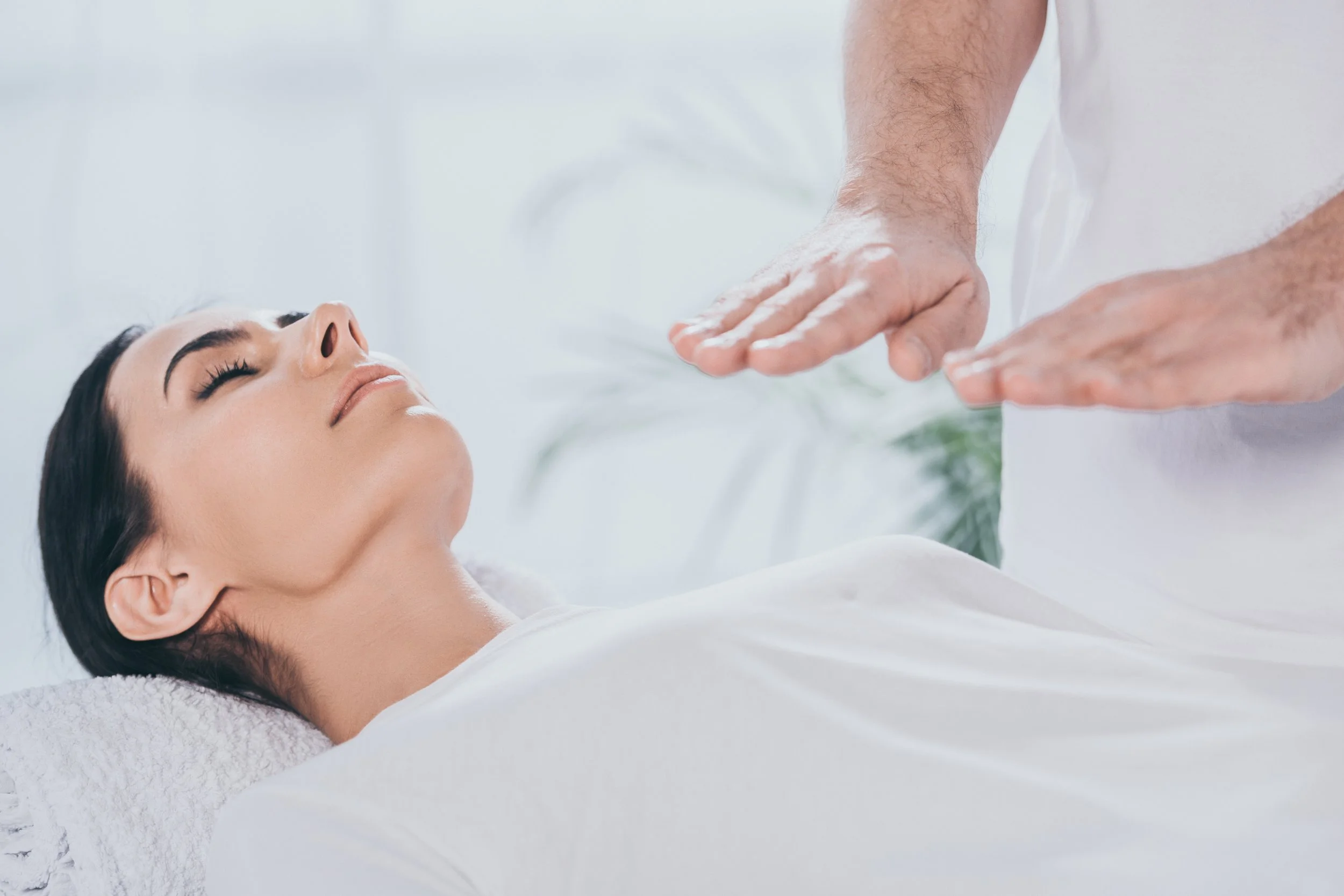 A woman lying down with her eyes closed, receiving a gentle hand massage from a person in a white uniform. The background is bright and minimalistic, suggesting a spa or wellness setting.