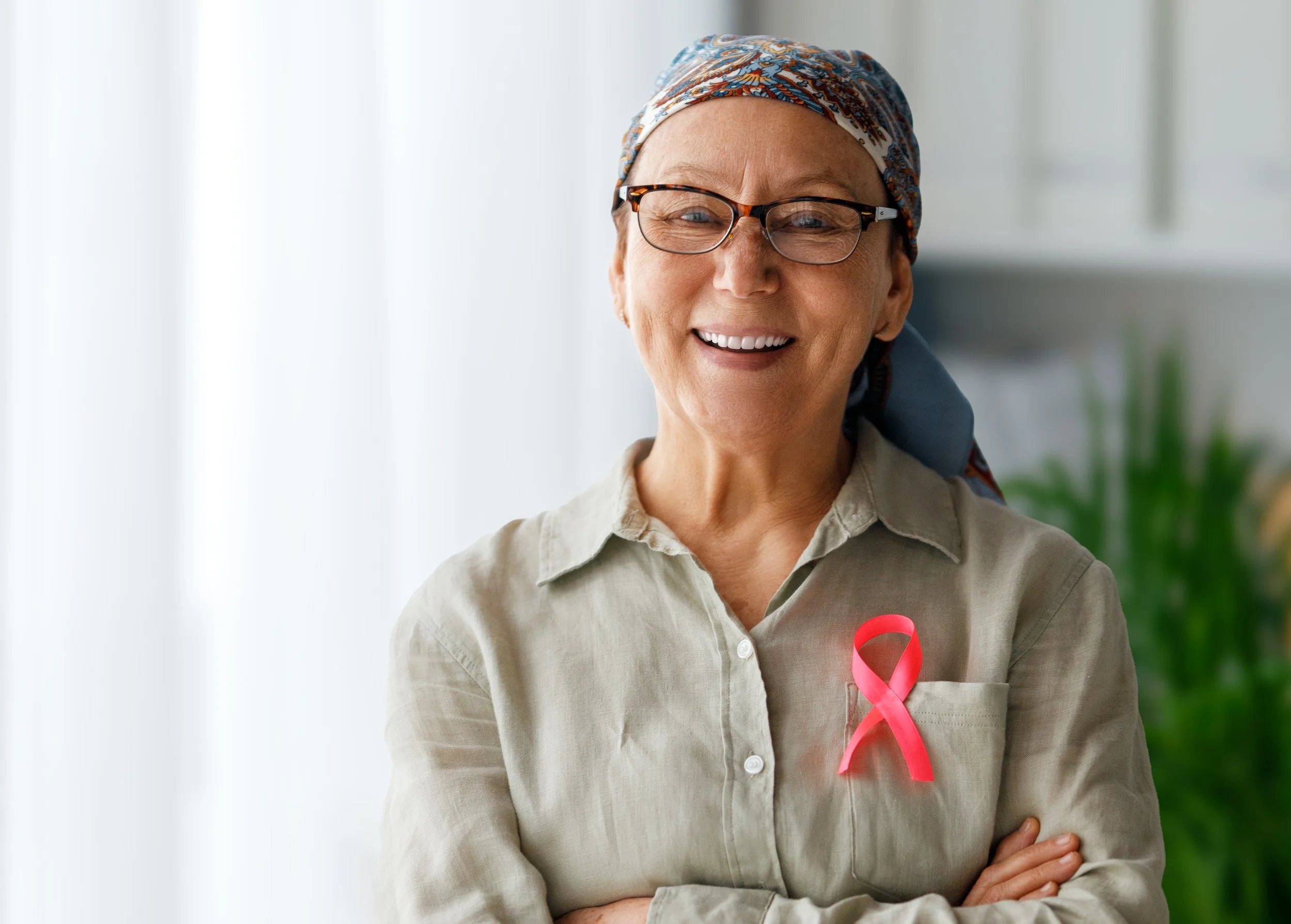 A smiling woman in glasses and a headscarf, wearing a beige shirt, with a red ribbon pinned to her chest, standing indoors with natural light and greenery in the background.