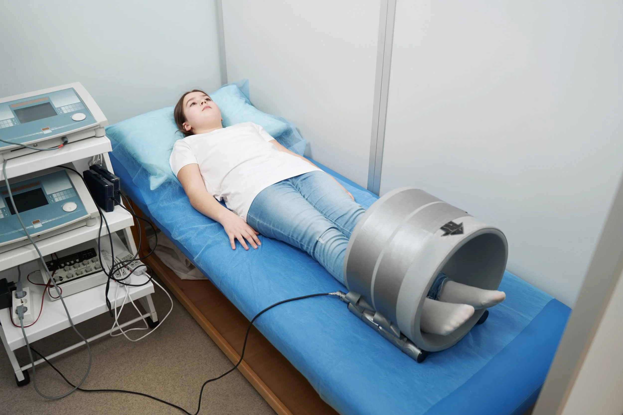 A young girl lying on a hospital bed with her leg in a medical device, connected to medical equipment, in a clinical room.