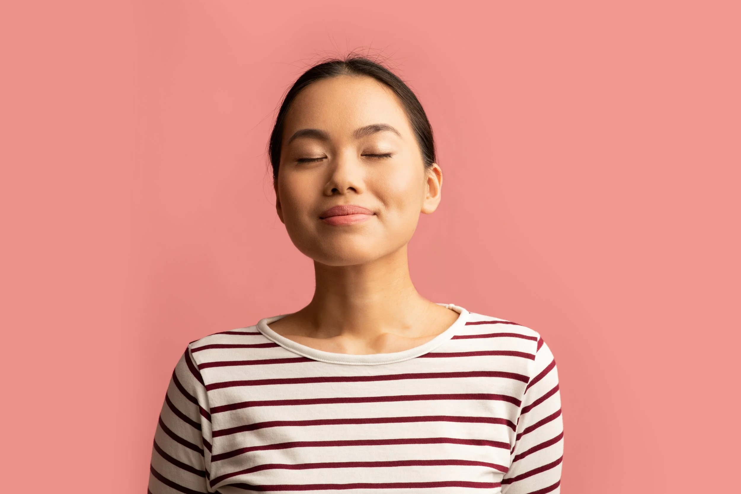 A young woman with closed eyes and a peaceful smile, wearing a striped shirt, standing against a pink background.