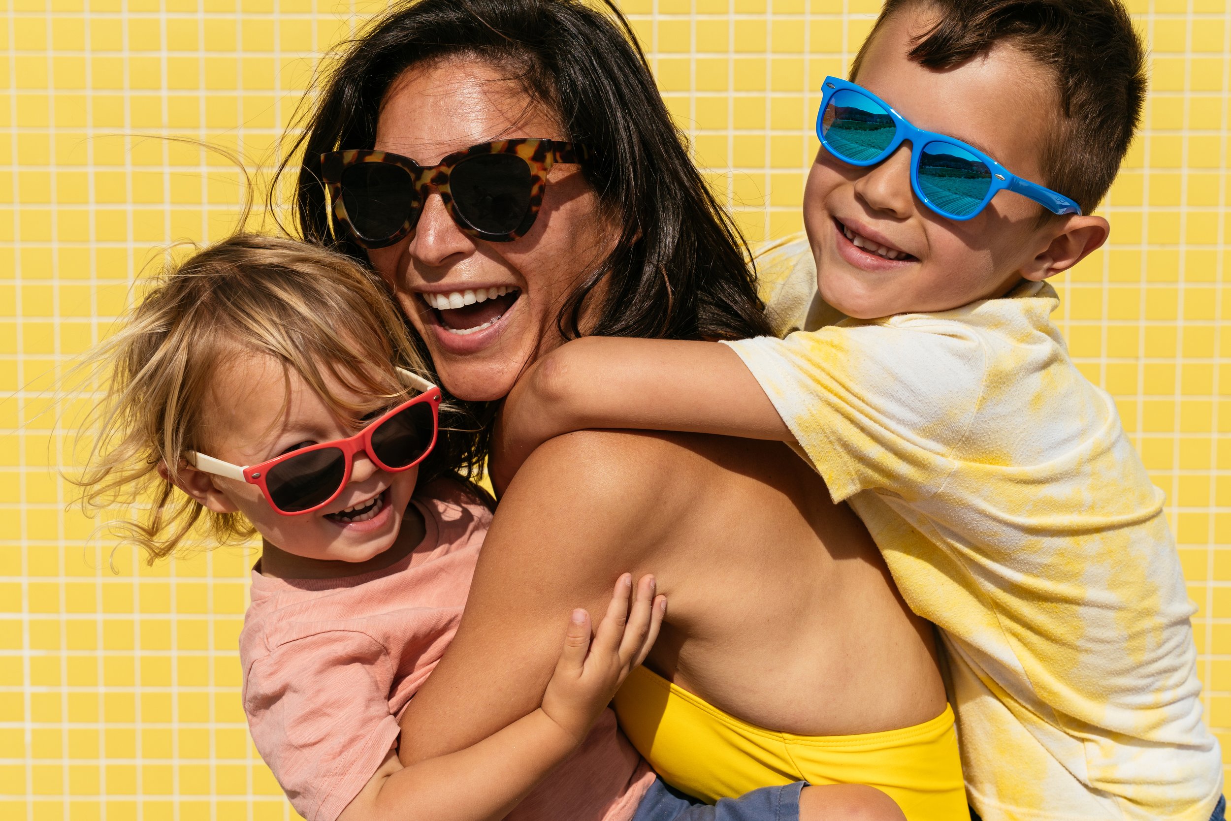 A woman with black hair and two children, a girl and a boy, all wearing colorful sunglasses, smiling and hugging outdoors in front of a yellow tiled wall.