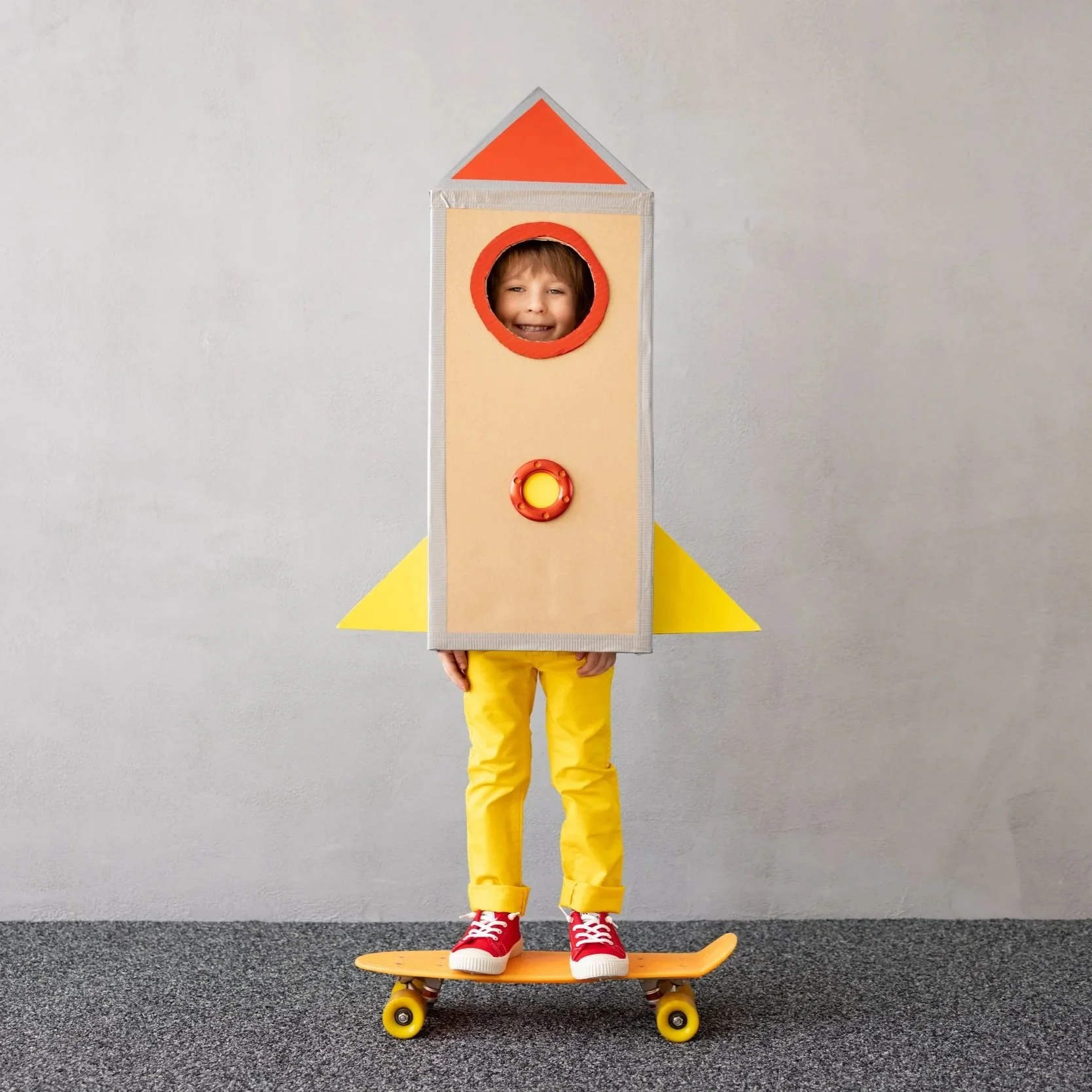 Child dressed as a rocket ship, standing on a skateboard, smiling through a round cutout in the costume, wearing red sneakers, yellow pants, and a cardboard rocket costume with yellow and orange accents, against a plain gray background.