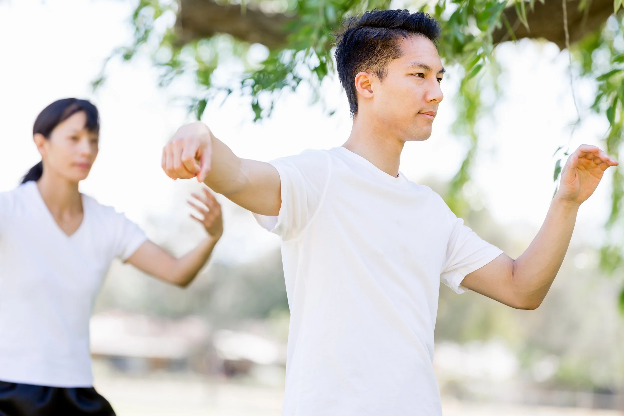 Two people practicing yoga outdoors with trees in the background. The man in the foreground has black hair, beige skin, and is wearing a white t-shirt. The woman in the background has black hair tied back, beige skin, and is also wearing a white t-shirt. Both are in a stretching pose with arms extended.