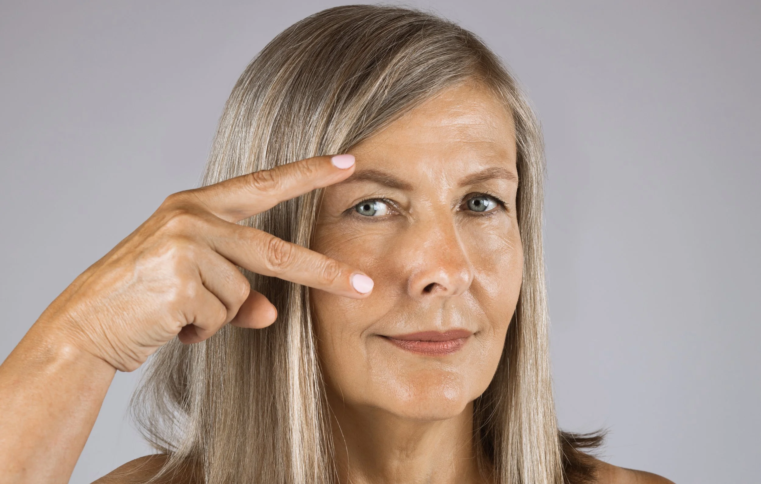 Close-up of a middle-aged woman with blonde hair and blue eyes, touching her forehead with her fingers, smiling softly against a plain gray background.