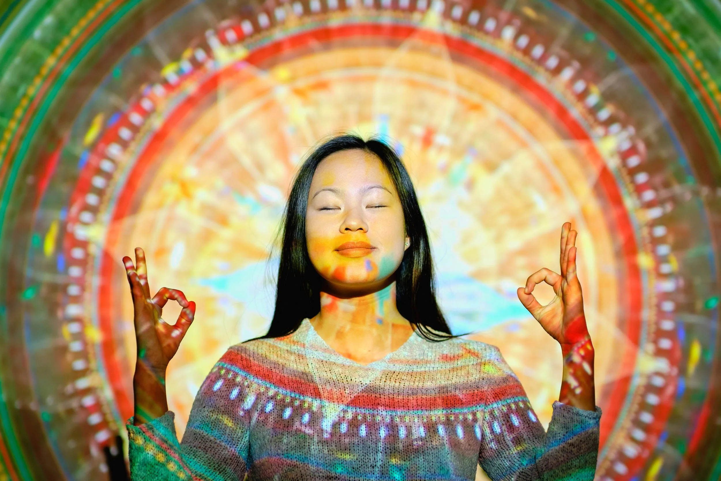 A woman with long dark hair making a meditation gesture with her hands in front of her chest, eyes closed, with a colorful, circular, rainbow-like light projection in the background.