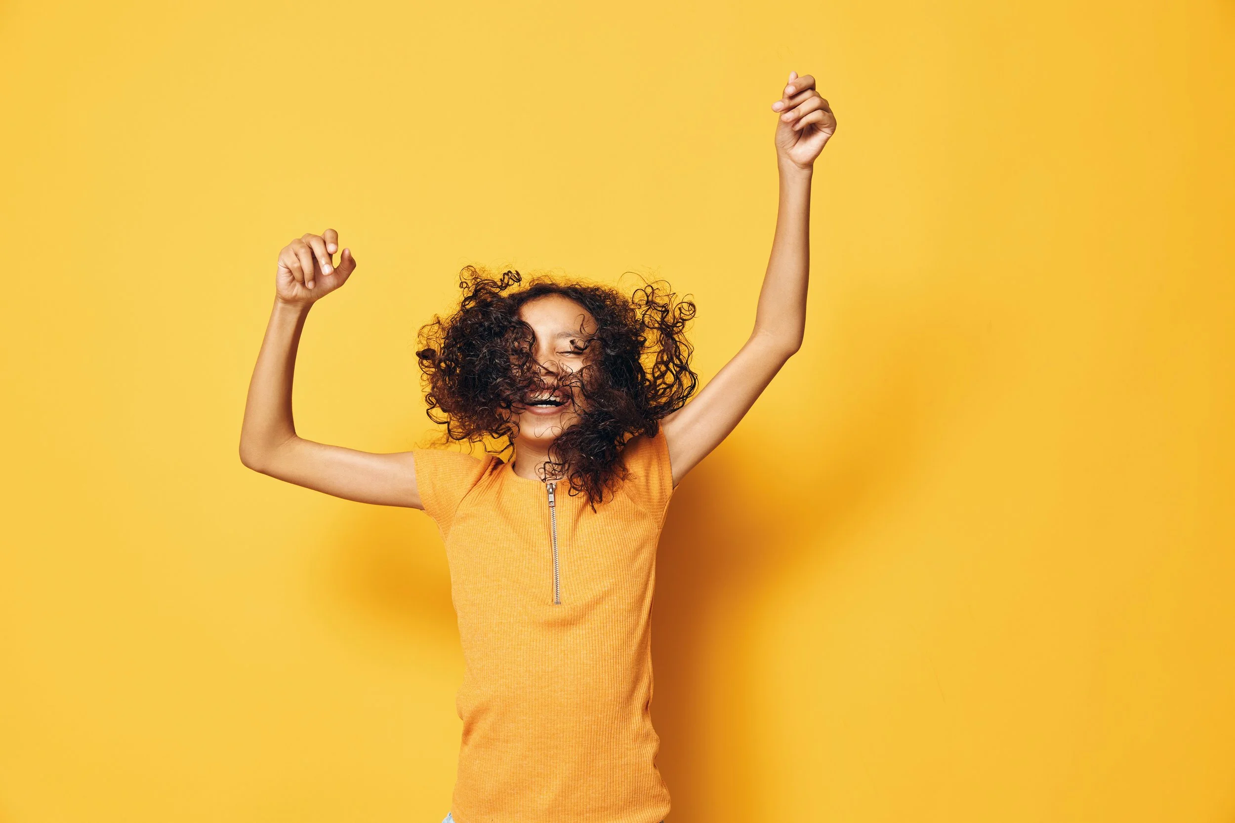 A person with curly hair wearing an orange top with a zipper smiling and dancing against a yellow background.