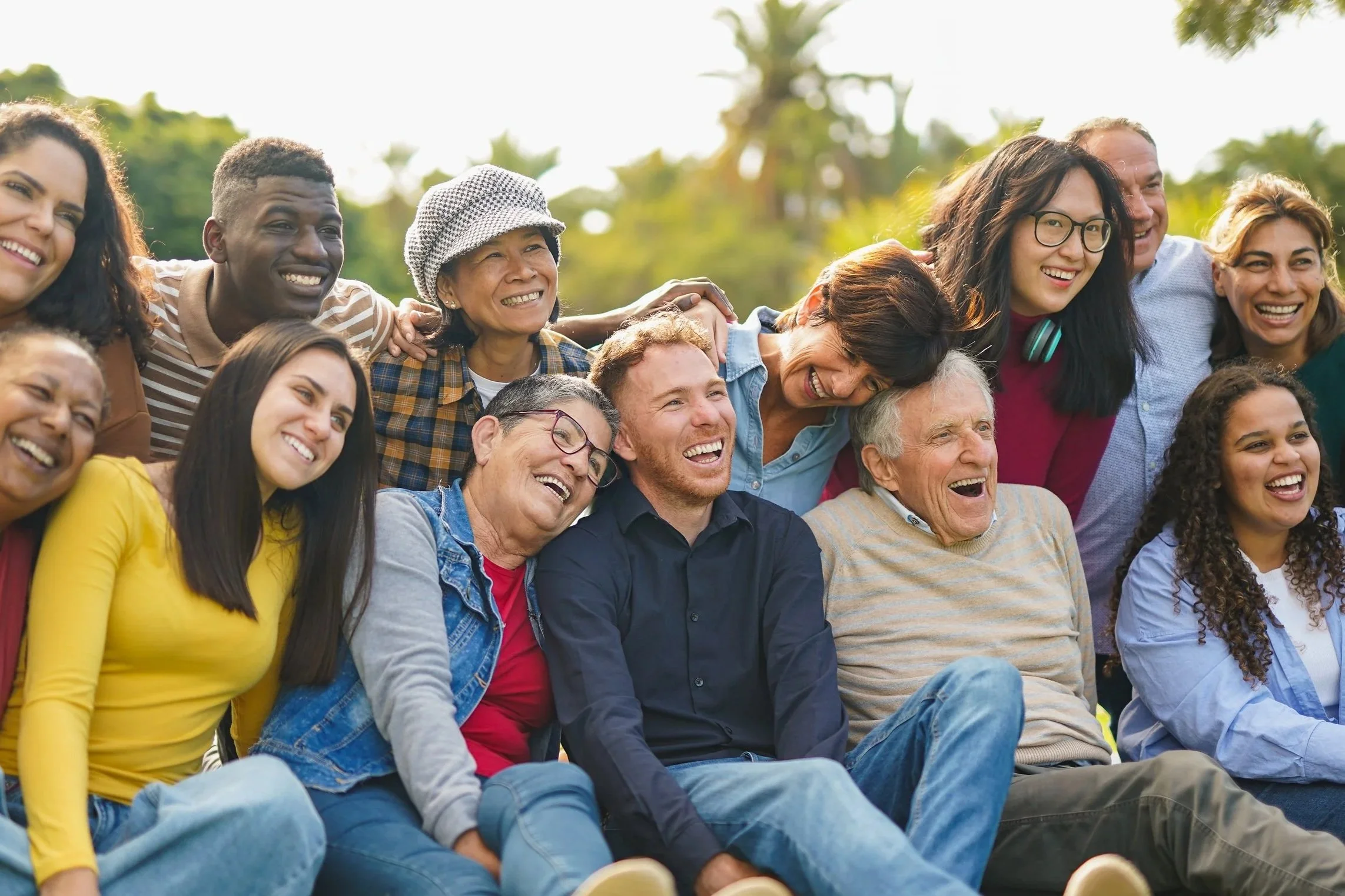 A diverse group of people sitting outdoors, smiling and posing for a photo with trees and clear sky in the background.