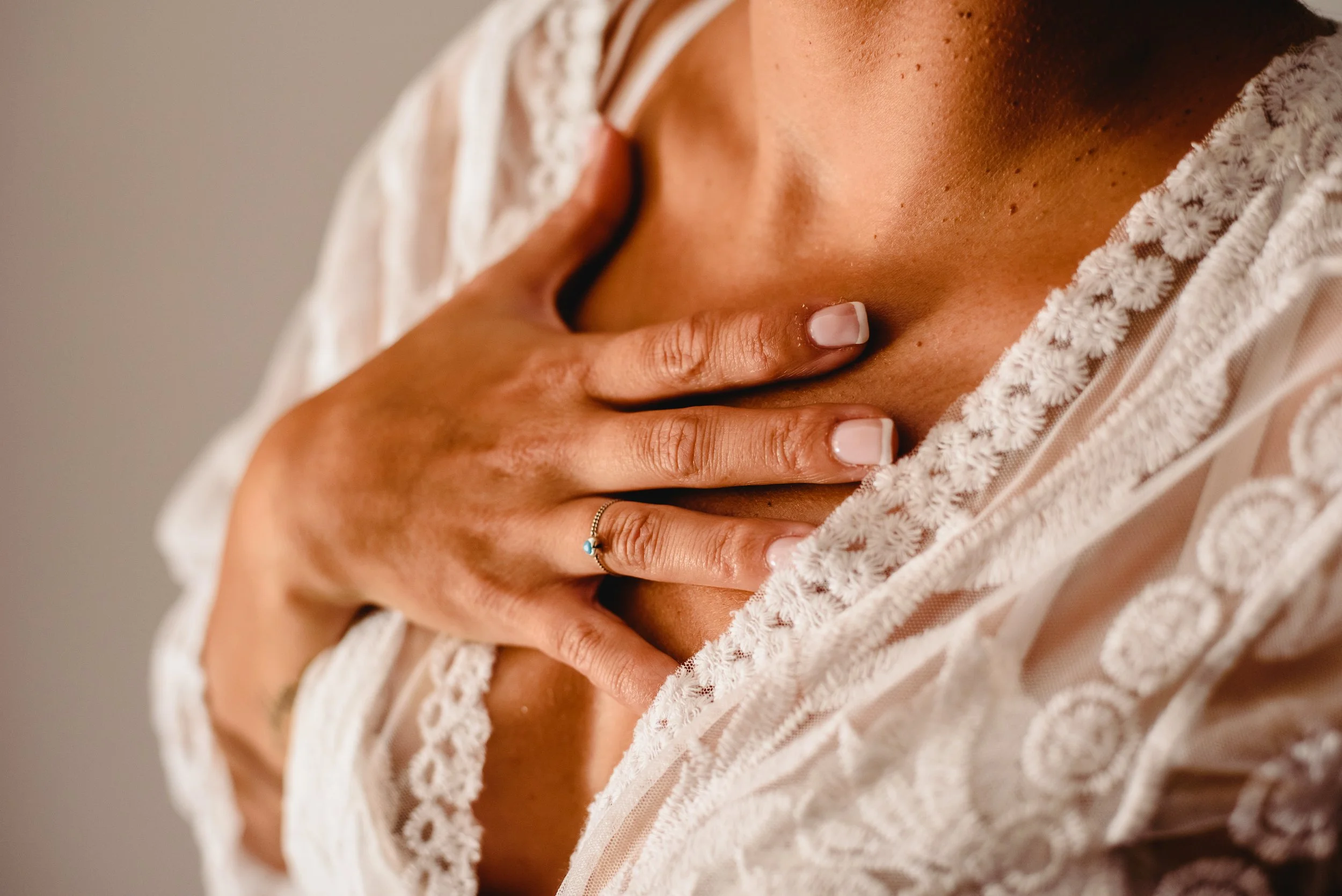 Close-up of a woman with tan skin, wearing a lace-trimmed shirt, holding her hand over her chest, showing her ring. The image focuses on her shoulder, neck, and collarbone area.