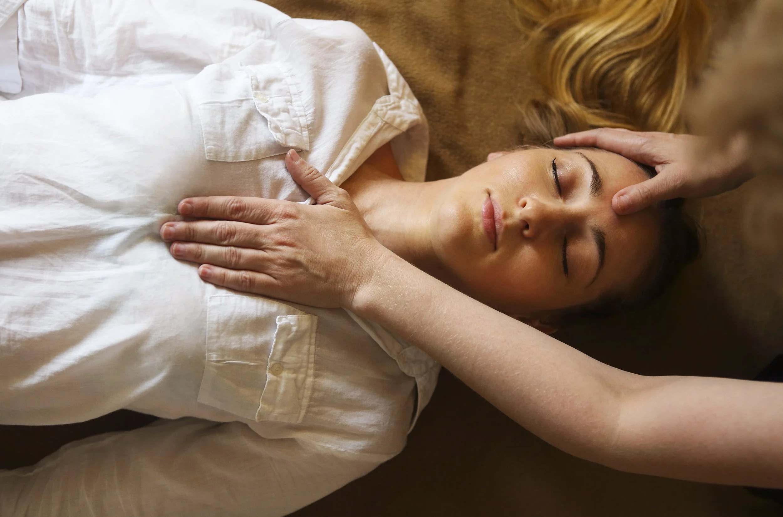 A woman receiving a head massage, lying on her back with eyes closed, wearing a white shirt.