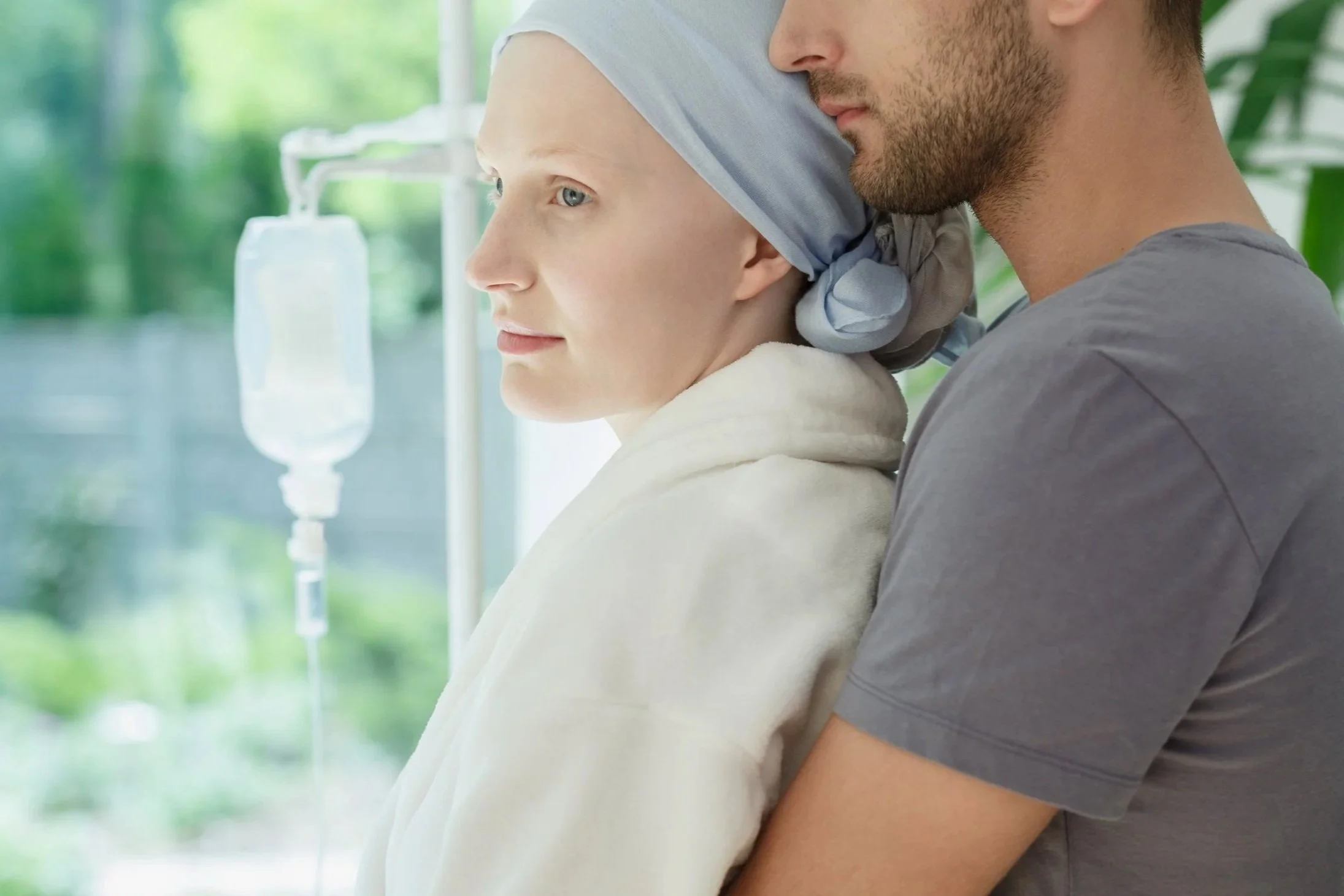 A woman with a headscarf and a man with a beard are close together at a healthcare setting, with IV fluid bags visible in the background.