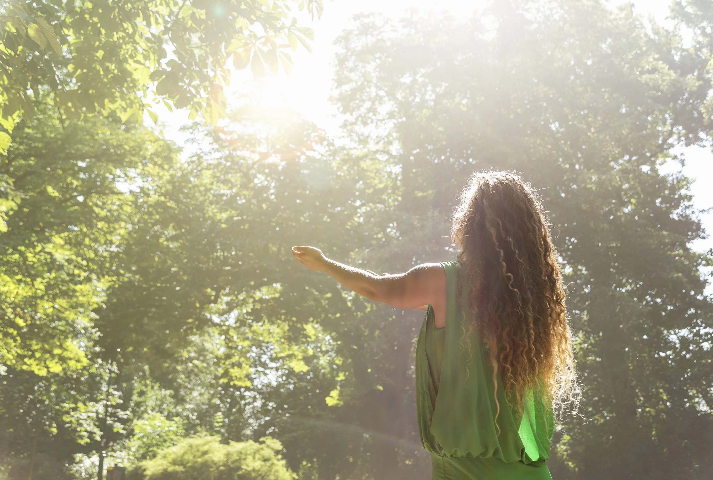 A person with long curly hair wearing a green dress standing outdoors among trees, with sunlight shining through the leaves.