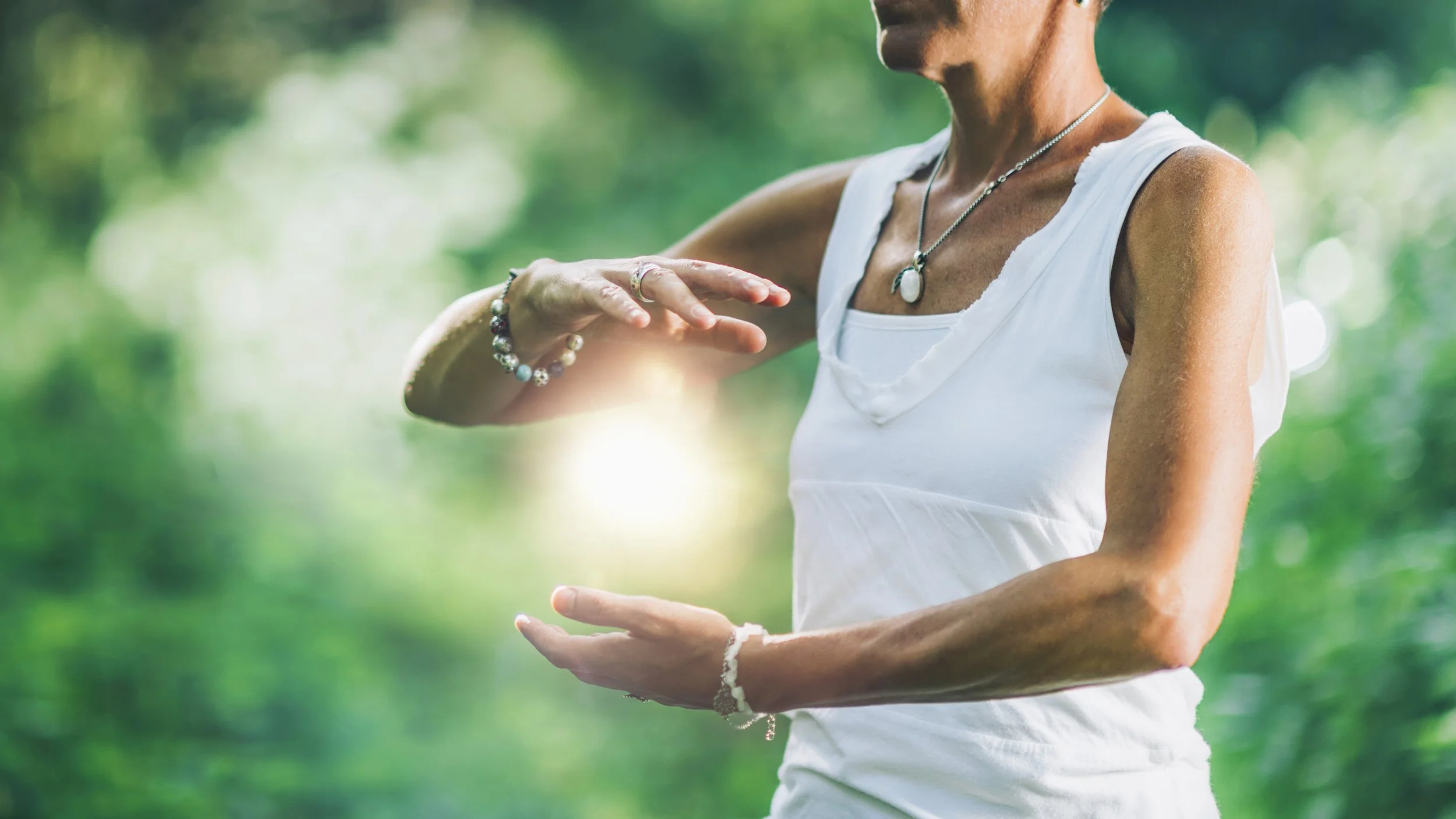 A woman outdoors with a blurred green background, wearing a white tank top and jewelry, looking at her wrist.