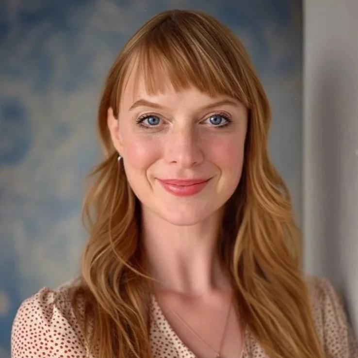 A woman with long red hair and blue eyes smiling at the camera, wearing a beige polka dot top and a necklace, with a blurred background.