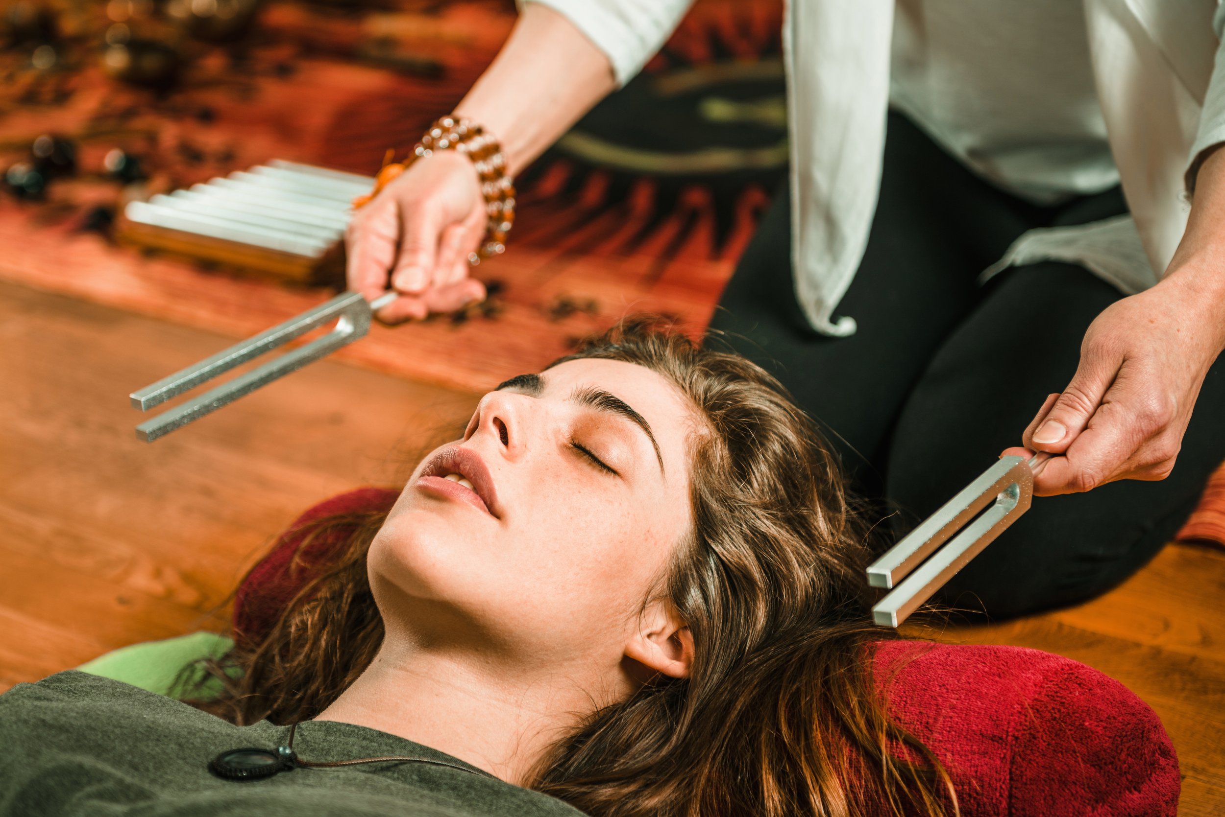 A woman lying on a wooden floor with her eyes closed while a person performs sound therapy with metal tuning forks on her forehead and chest.