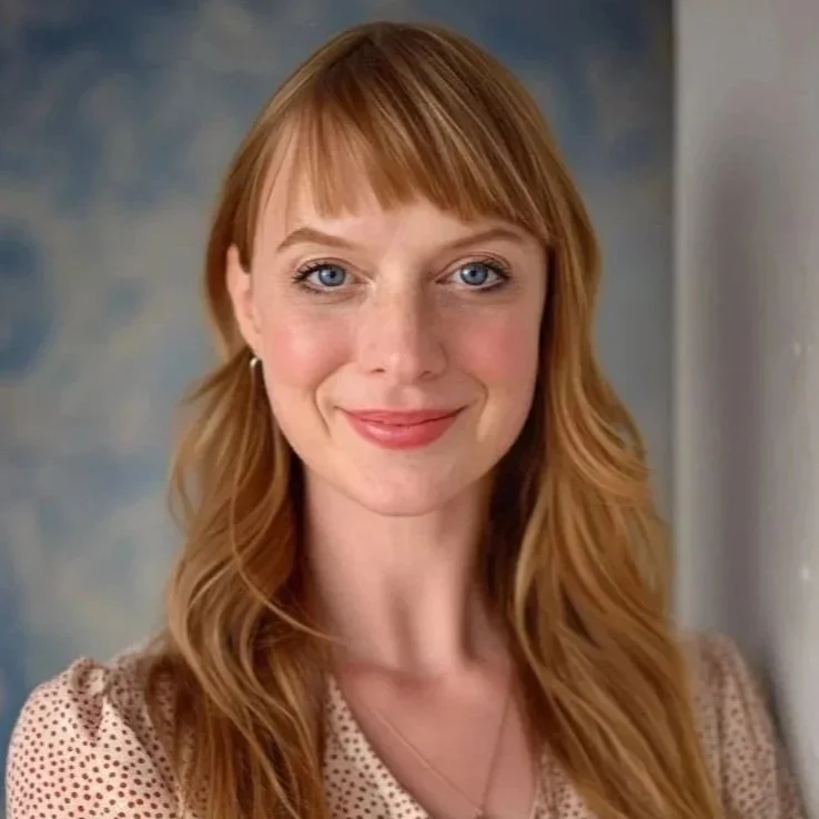 A woman with red hair, blue eyes, and fair skin smiling at the camera, wearing a light-colored polka dot top and a delicate necklace, with a soft blue and gray background.