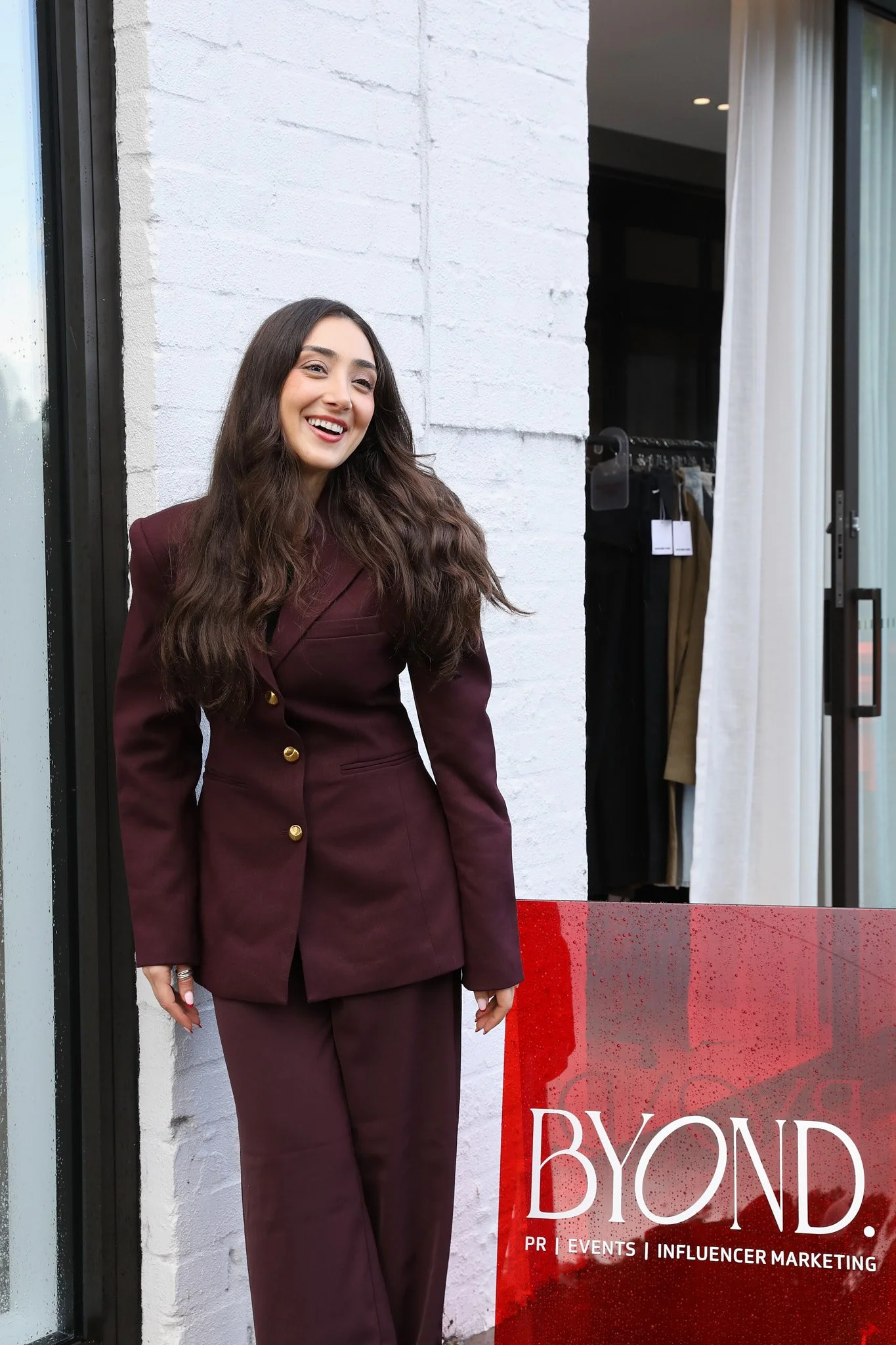 Pardis Mahdavi, founder and director of BYOND, a Sydney PR, events and influencer marketing agency, smiling in a burgundy suit beside the red BYOND sign.