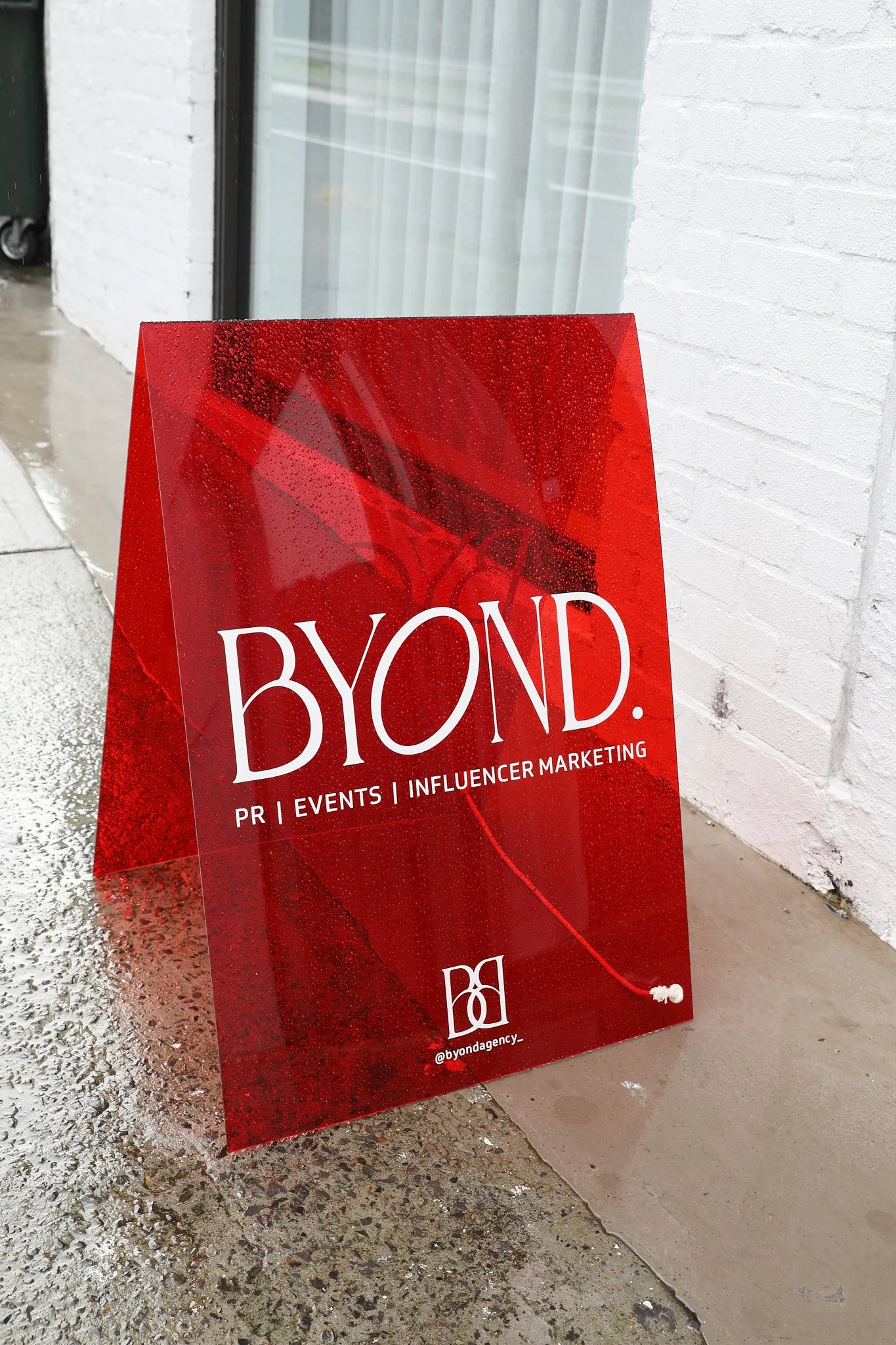 Red BYOND agency sign reading PR, Events and Influencer Marketing, with the handle @byondagency_, outside a white brick building.
