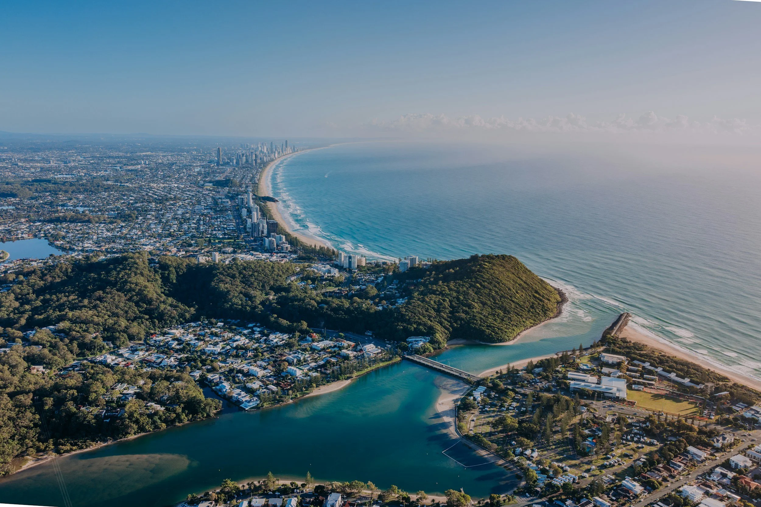 An aerial view of a coastal city with high-rise buildings along the shoreline, a long sandy beach, a river with a bridge, and surrounding green hills under a clear blue sky.