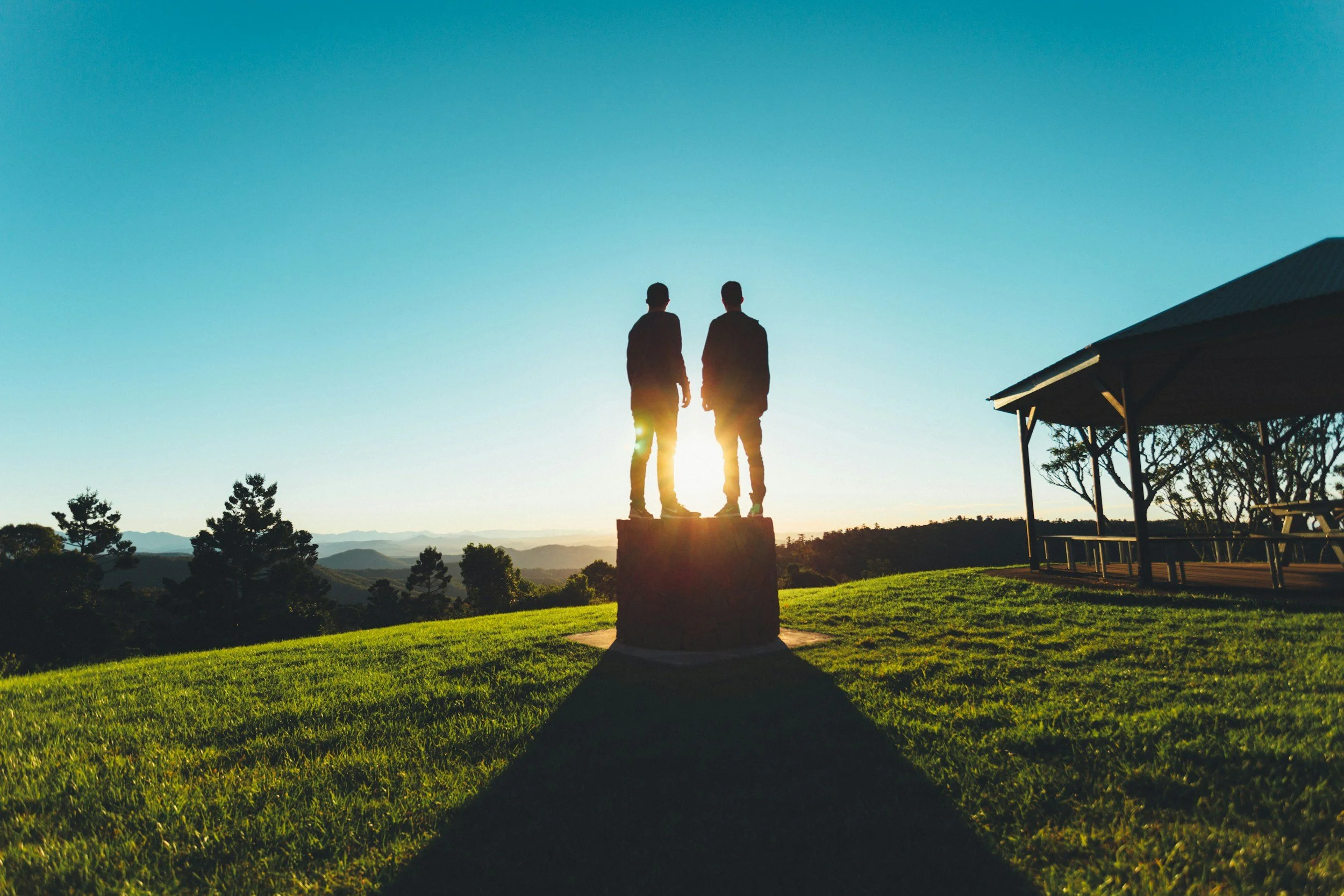 Two people standing on a pedestal outdoors at sunset, with a landscape and mountains in the background; trees and a pavilion are visible in the grassy area.