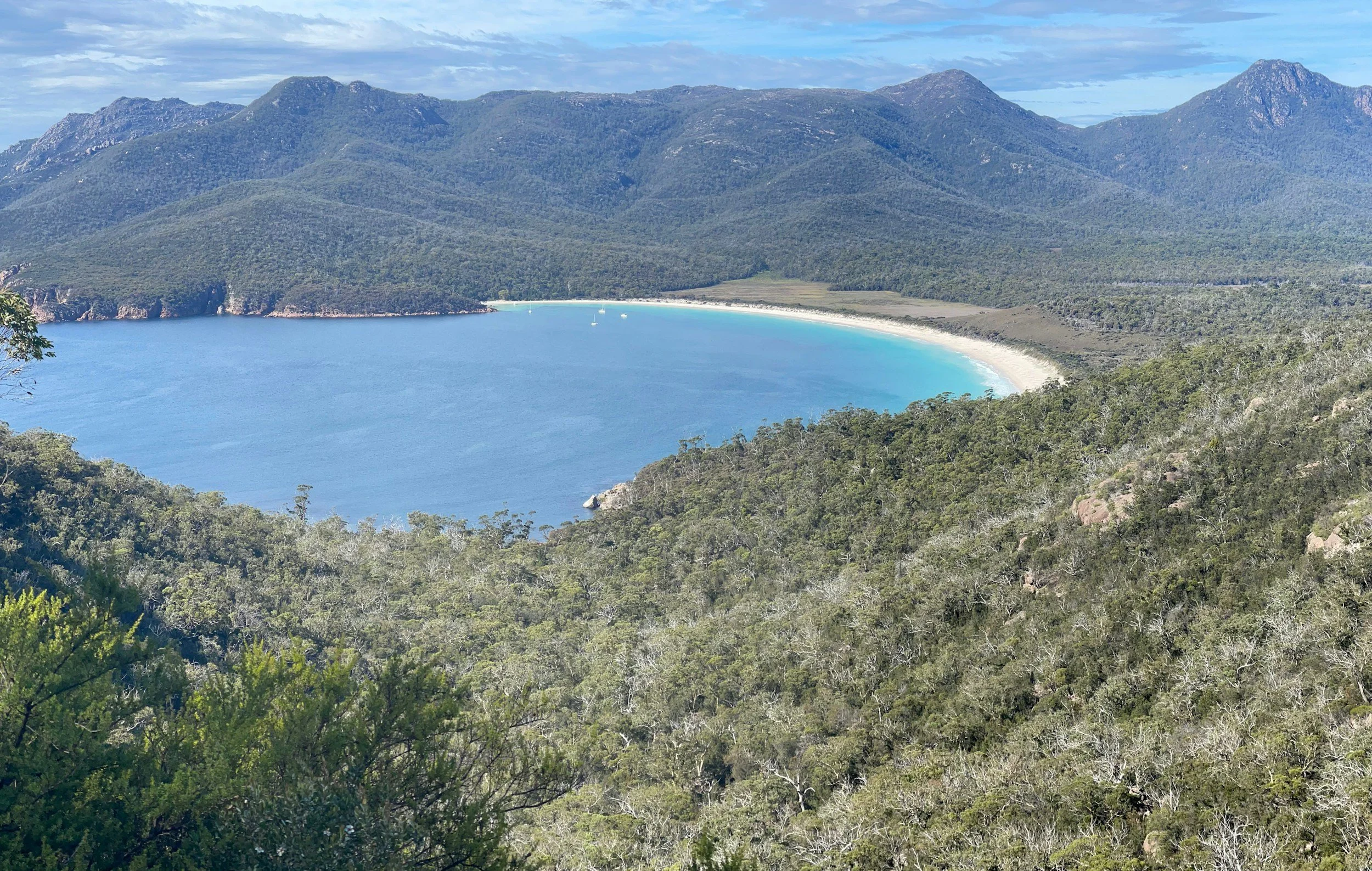 Scenic view of a bay surrounded by green mountains with a white sandy beach and boats on the water.