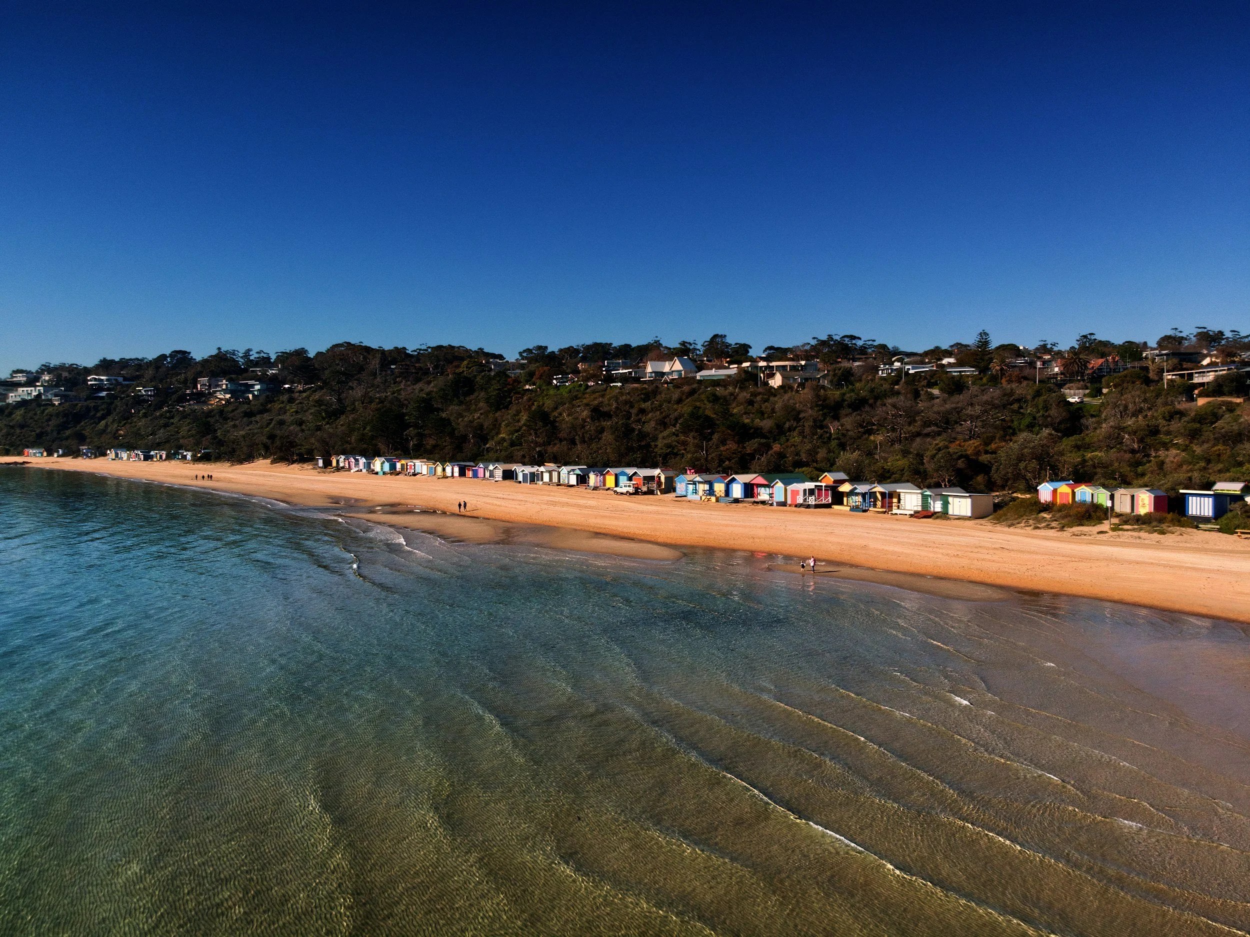 A beach with colorful beach huts along the shoreline, green trees on a hill, residential houses on the hilltop, and clear blue sky overhead.