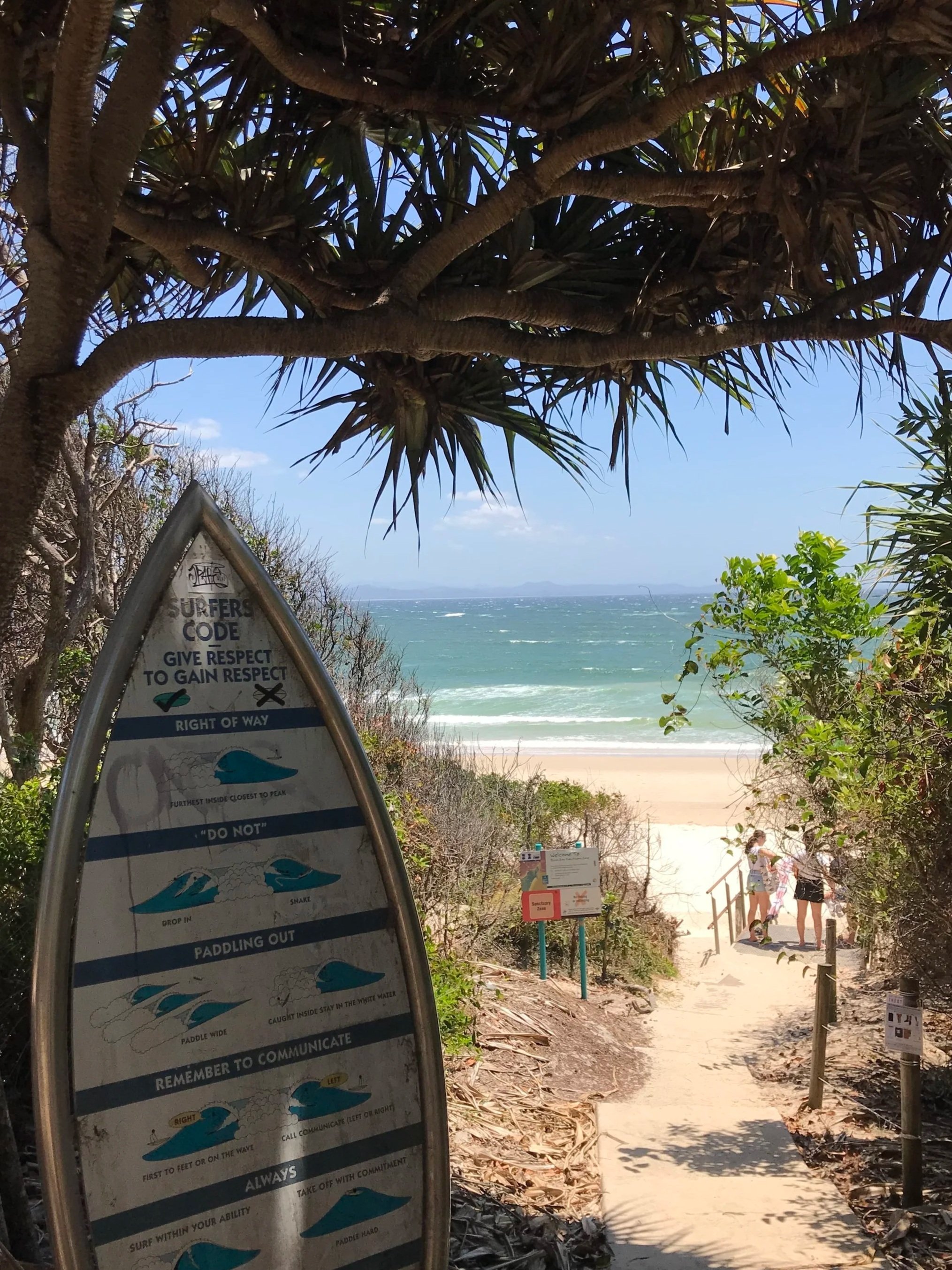 A view of a beach pathway leading to the ocean, with a signpost on the left listing surf safety rules. The pathway is surrounded by greenery and trees, with a few people walking towards the beach. The ocean and a distant landmass are visible in the background.
