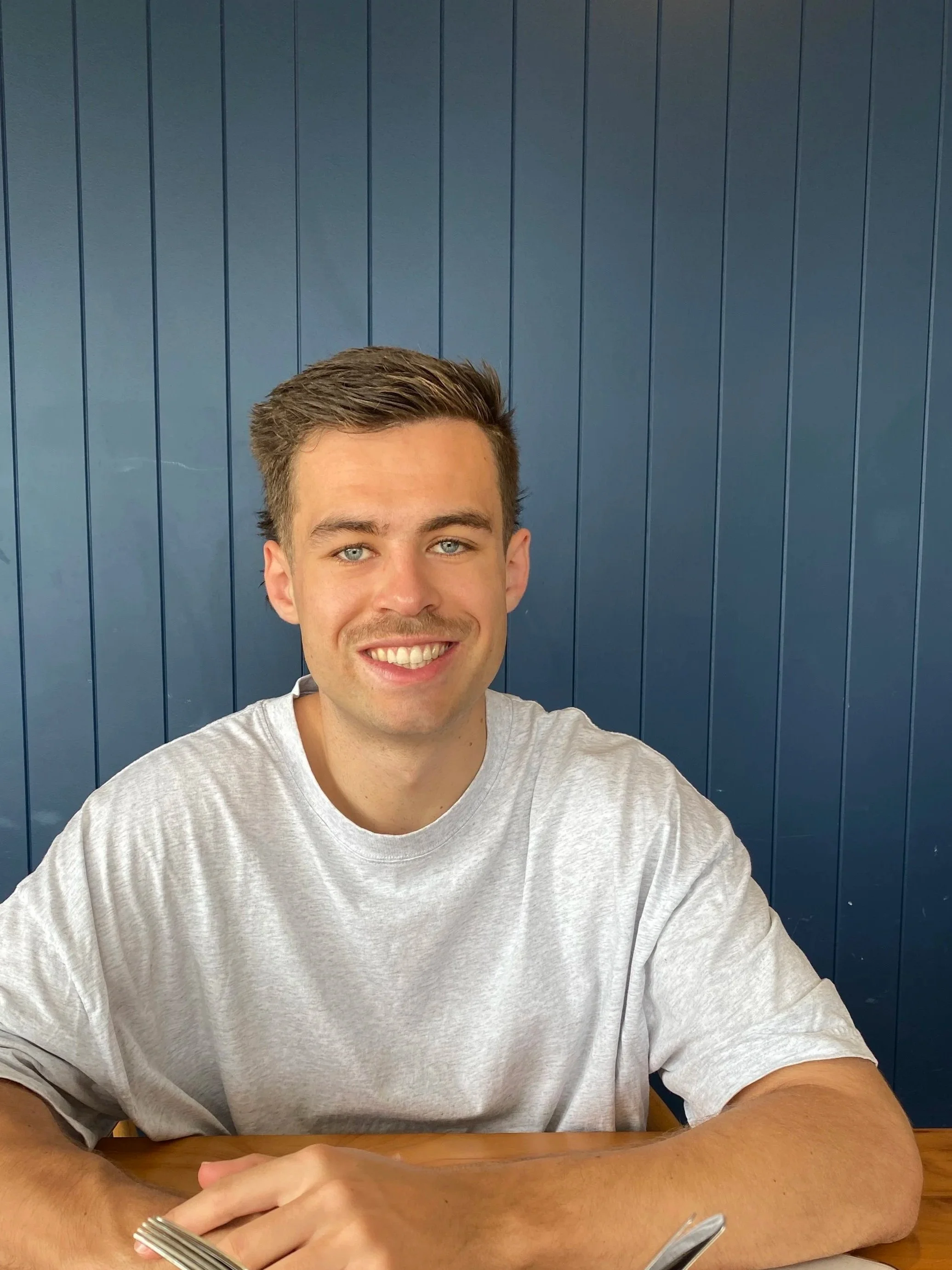 A young man with short brown hair and blue eyes smiling, sitting at a table with a silver fork in his hand, against a blue wooden panel background.