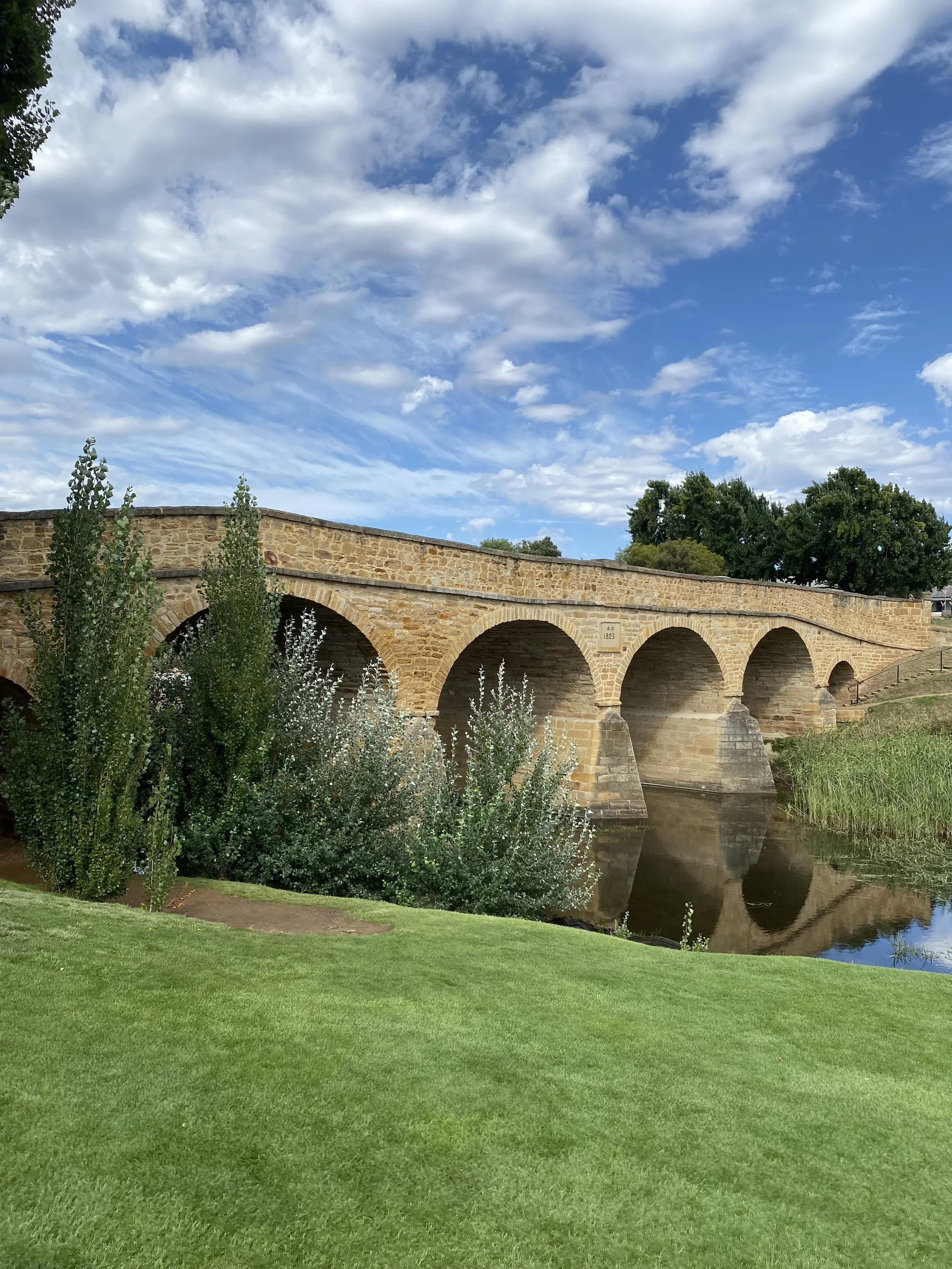 A stone bridge with arches spans over a calm river, surrounded by lush green grass and trees, under a blue sky with scattered white clouds.