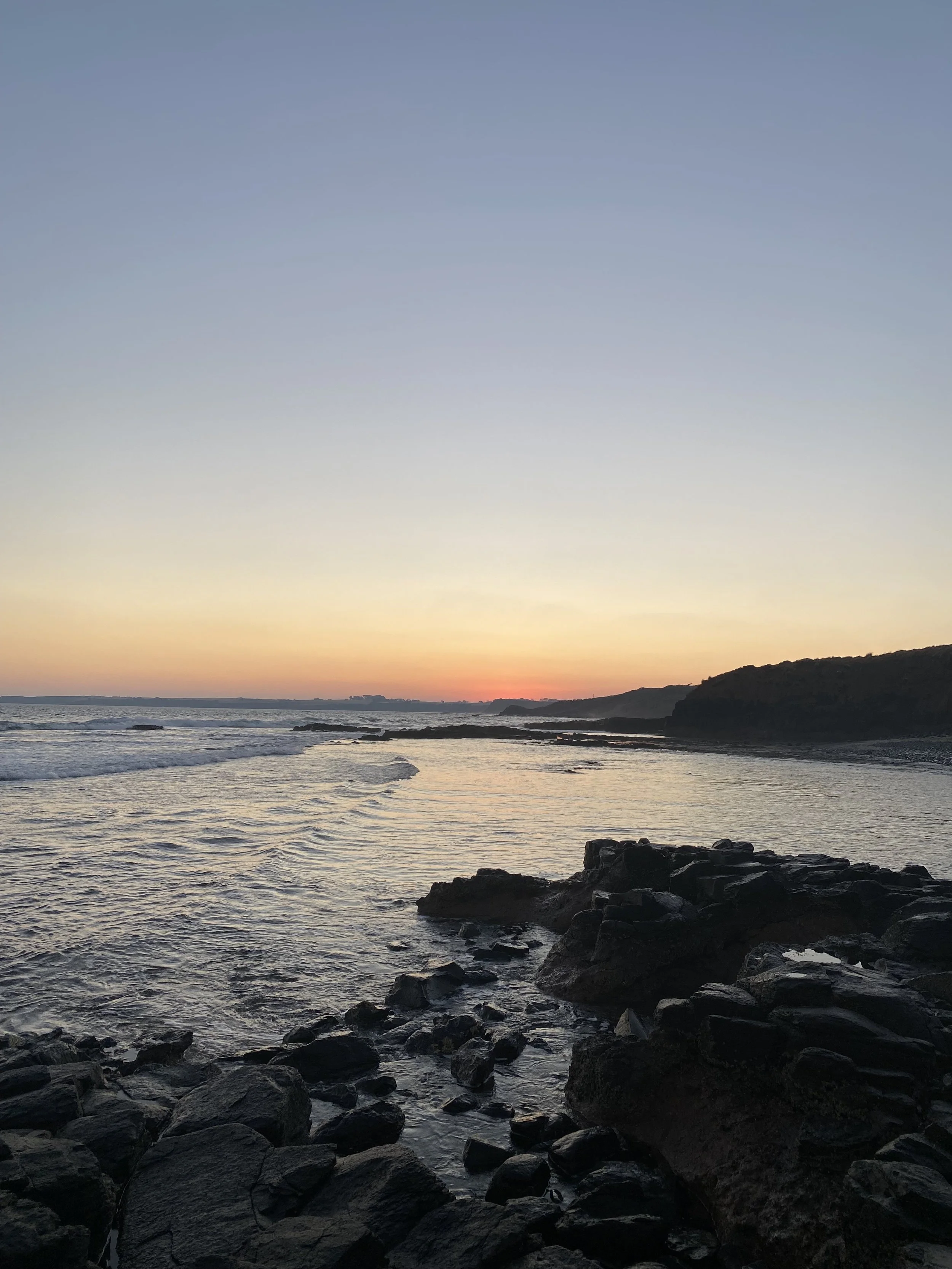 View of a seashore during sunset with rocks in the foreground, calm waves, and hills on the horizon under a partly cloudy sky.