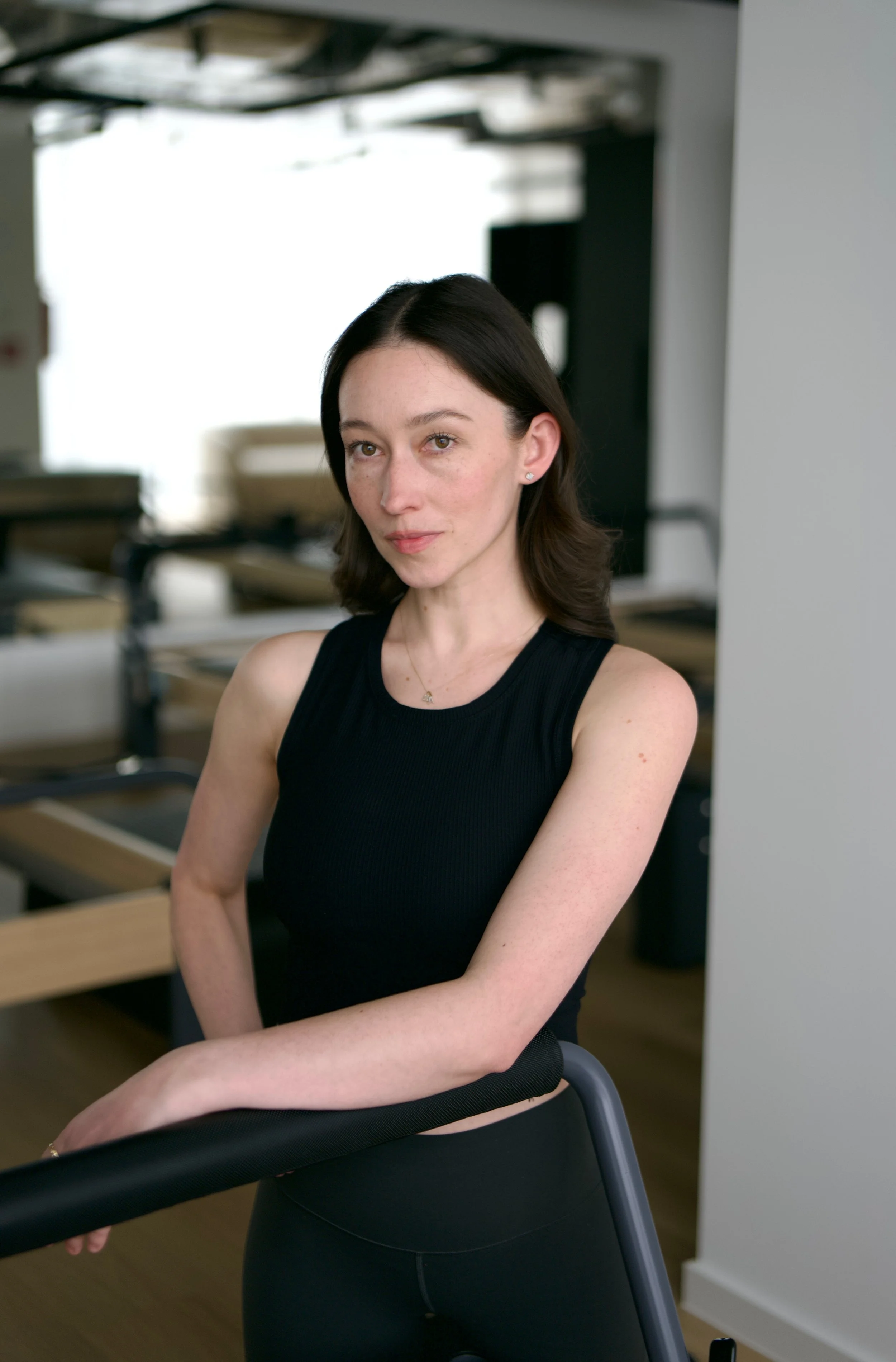A young woman with shoulder-length dark hair, wearing a black sleeveless top and black leggings, is sitting on a chair in a bright room with wooden floors and fitness equipment in the background.