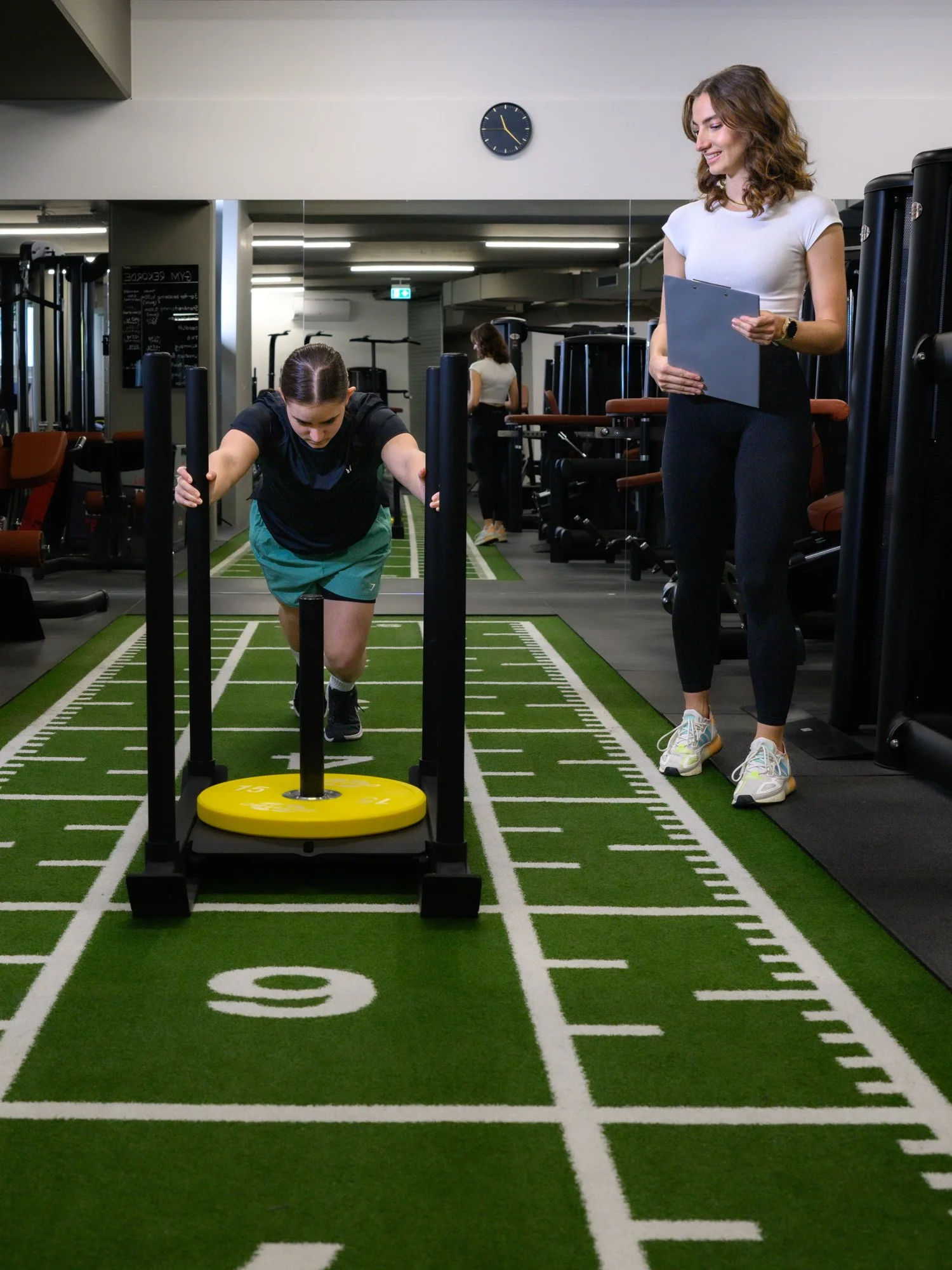 Eine Frau schiebt einen Schlitten mit einem gelben Gewicht, während die Physiotherapeutin mit Notizblock zuschaut.