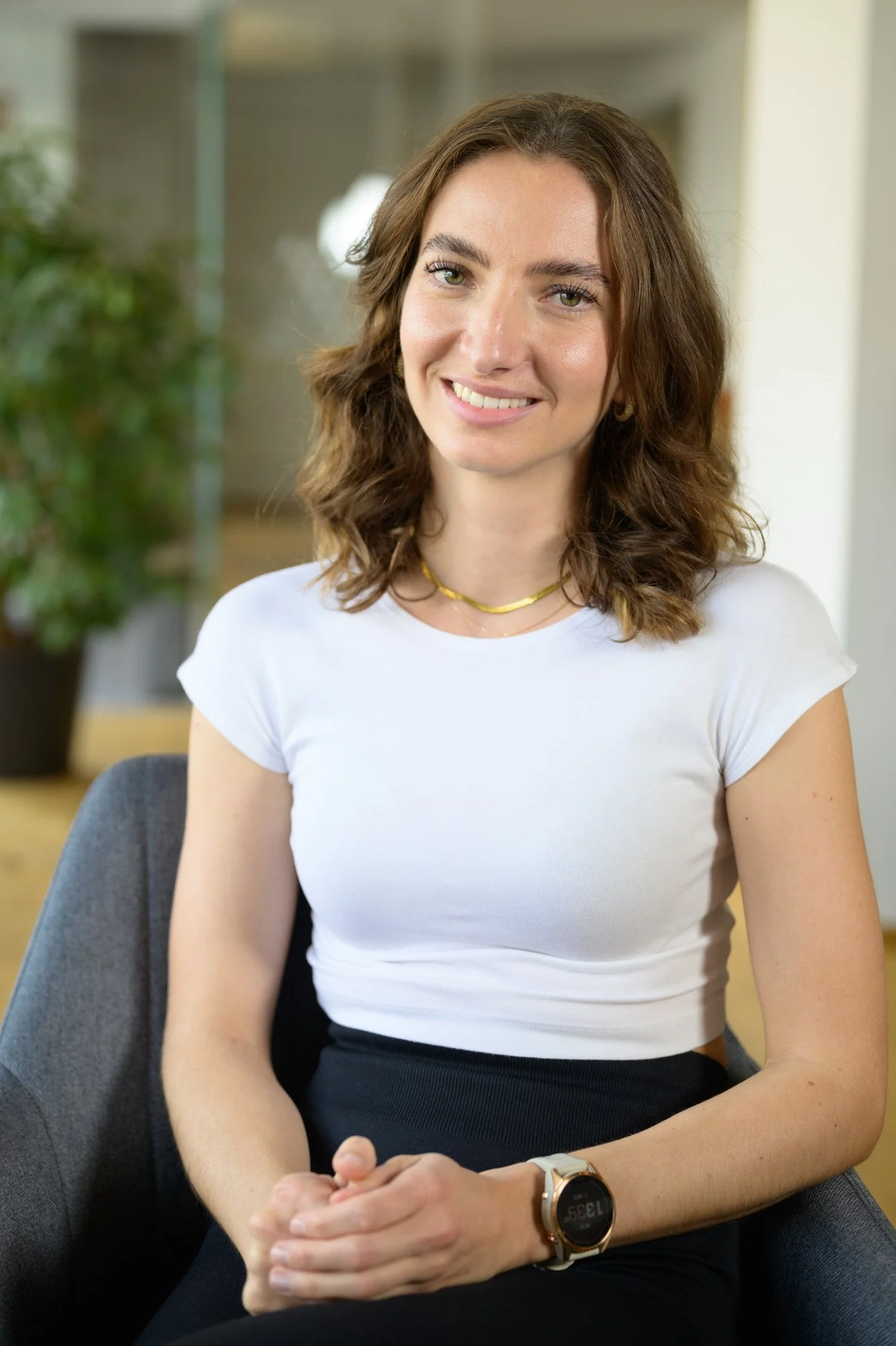 Eine lächelnde Frau mit braunen, lockigen Haaren sitzt in einem modernen Büro. Sie trägt ein weißes T-Shirt, eine goldene Halskette und eine Smartwatch.