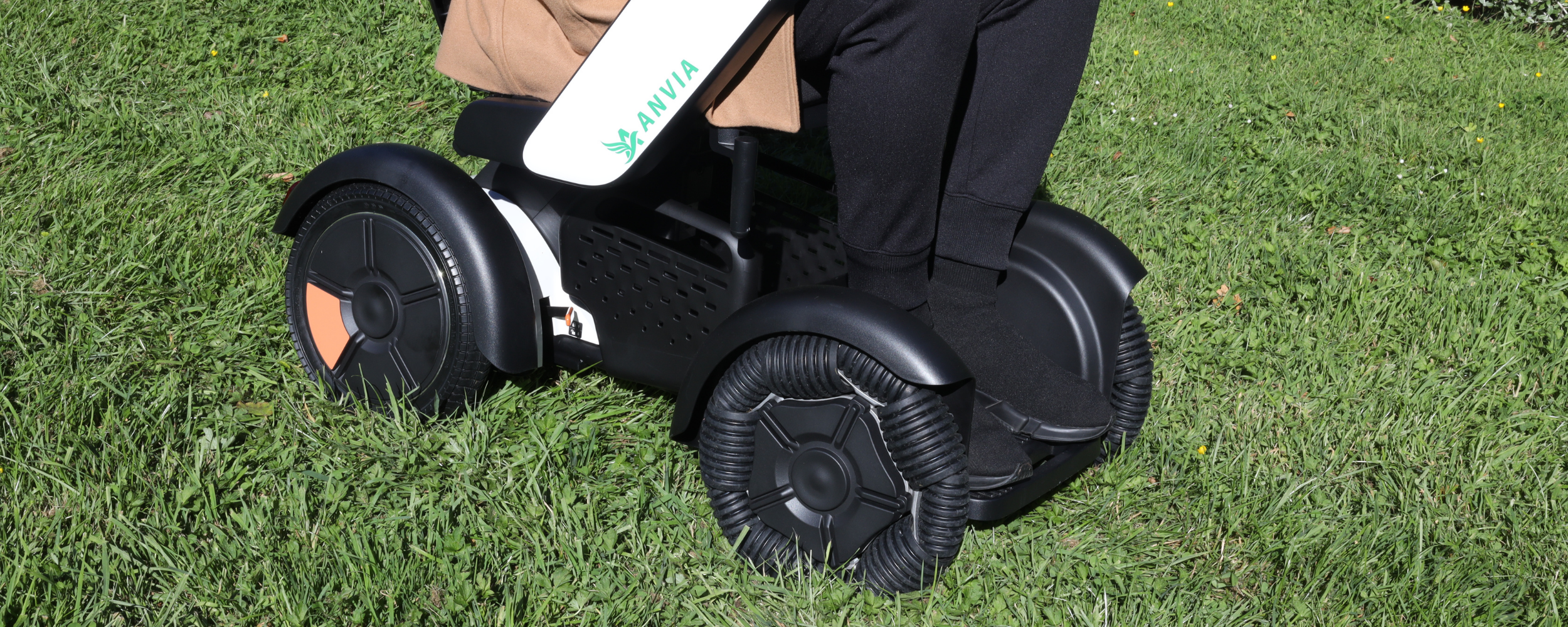 Close-up of an electric stroller with black and white colors, large black wheels, and a person wearing black pants and a brown jacket sitting on it, on a grassy field.