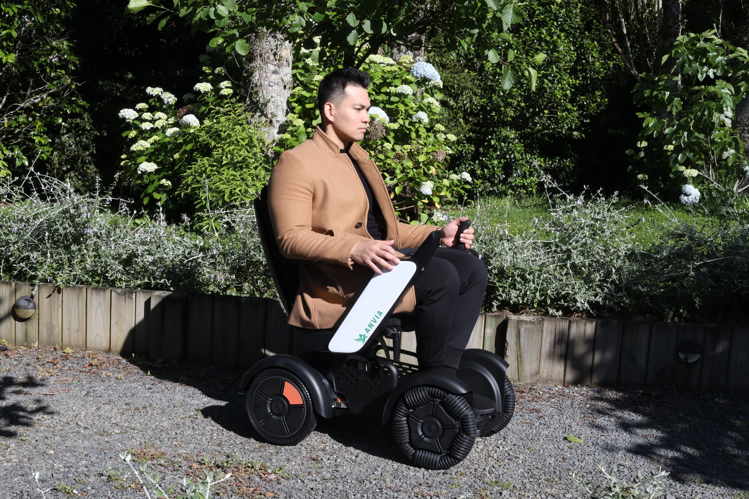 Man sitting outdoors on a branded electric mobility device with wheels and a handle, surrounded by lush greenery and flowers.