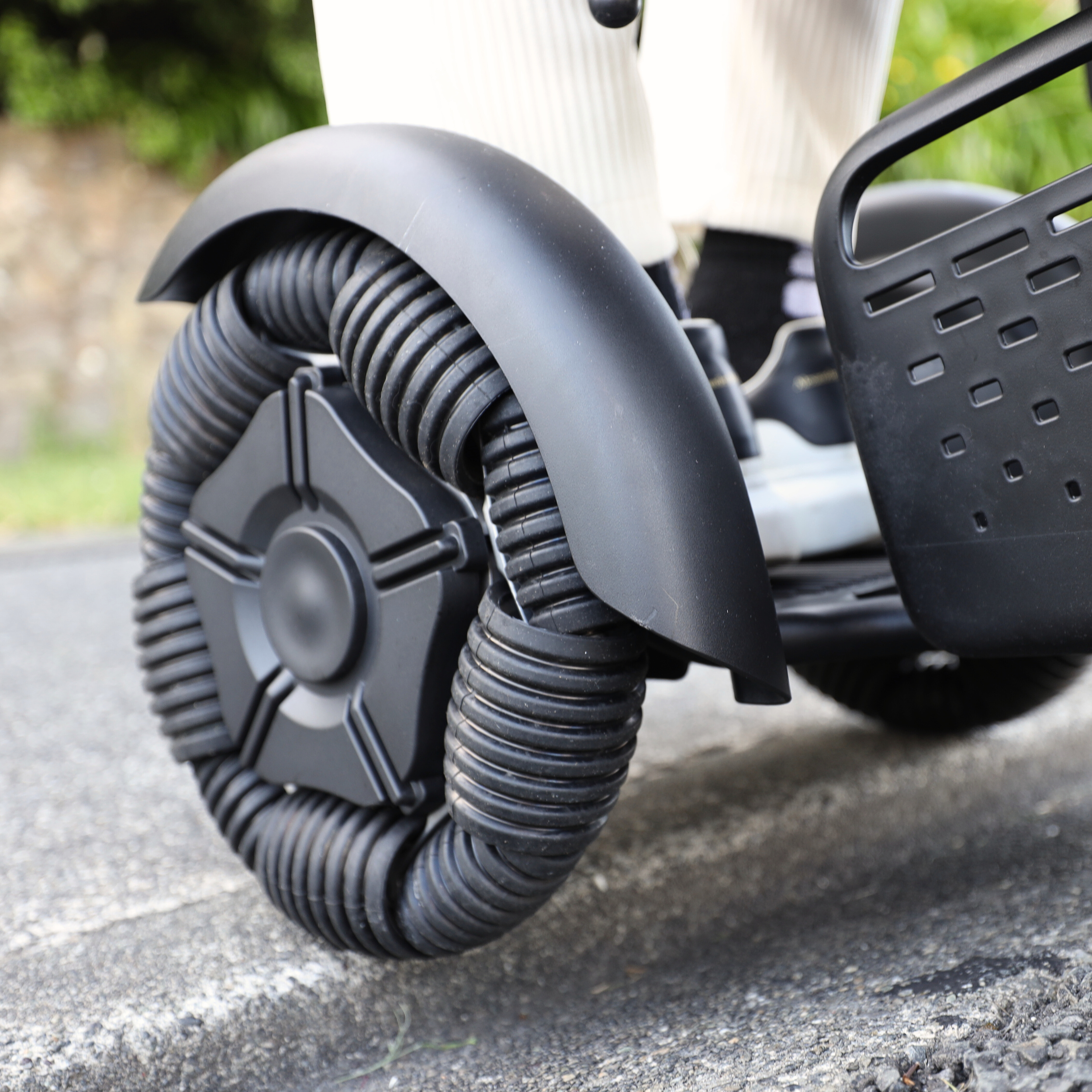 Close-up of a black, futuristic-looking, one-wheeled robot with large treads and a black seat, parked on a paved surface with greenery in the background.