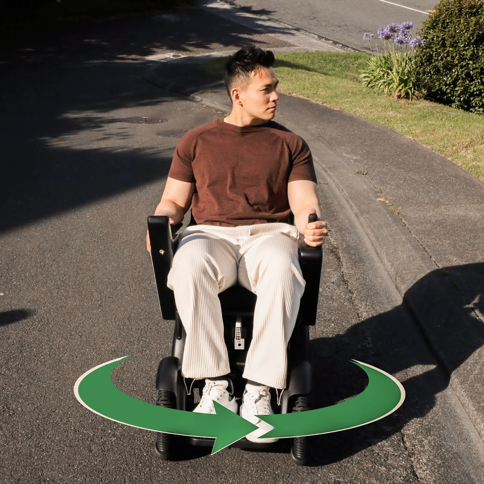 A young man in a brown t-shirt and white pants sitting in a motorized wheelchair outdoors on a sidewalk, with a green arrow indicating a swiveling rotation of the wheelchair into a different position.