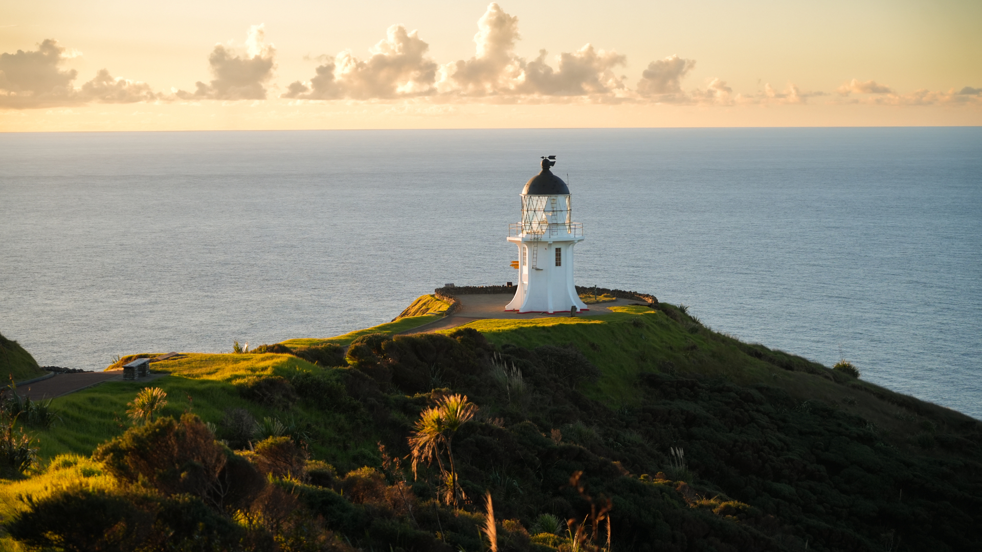A lighthouse on a grassy hill overlooking the ocean at sunset, with clouds in the sky.
