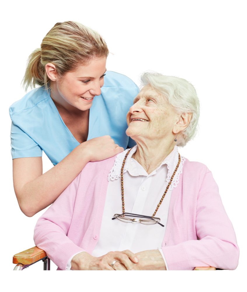 A young female healthcare worker smiling and leaning towards an elderly woman sitting in a wheelchair, who is also smiling and looking at her, wearing a pink cardigan and glasses hanging from her neck.