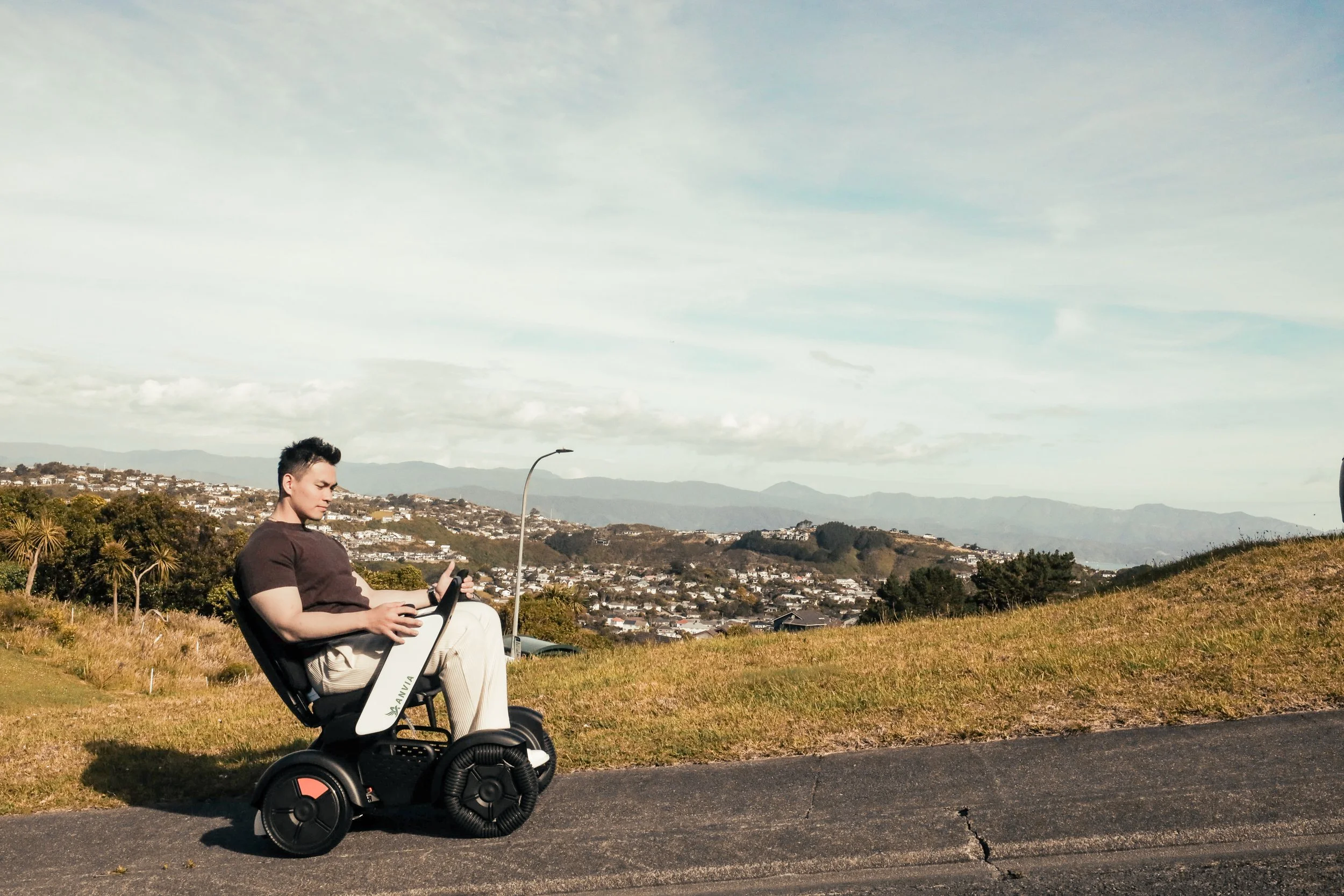 A man sitting on a white and black motorized wheelchair on a paved path outdoors, with grassy hills and a cityscape with houses and trees in the background, under a partly cloudy sky.