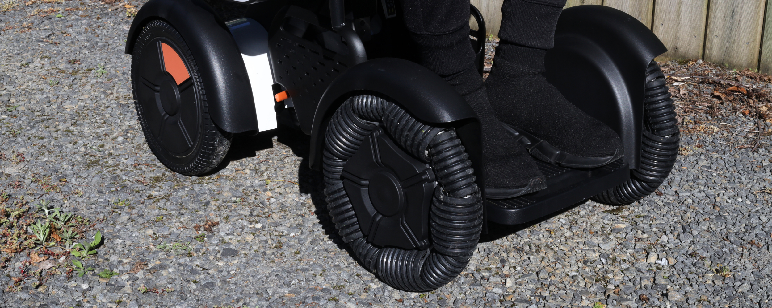 Close-up of the front and rear wheels of a motorized wheelchair on gravel, with a wooden fence in the background.