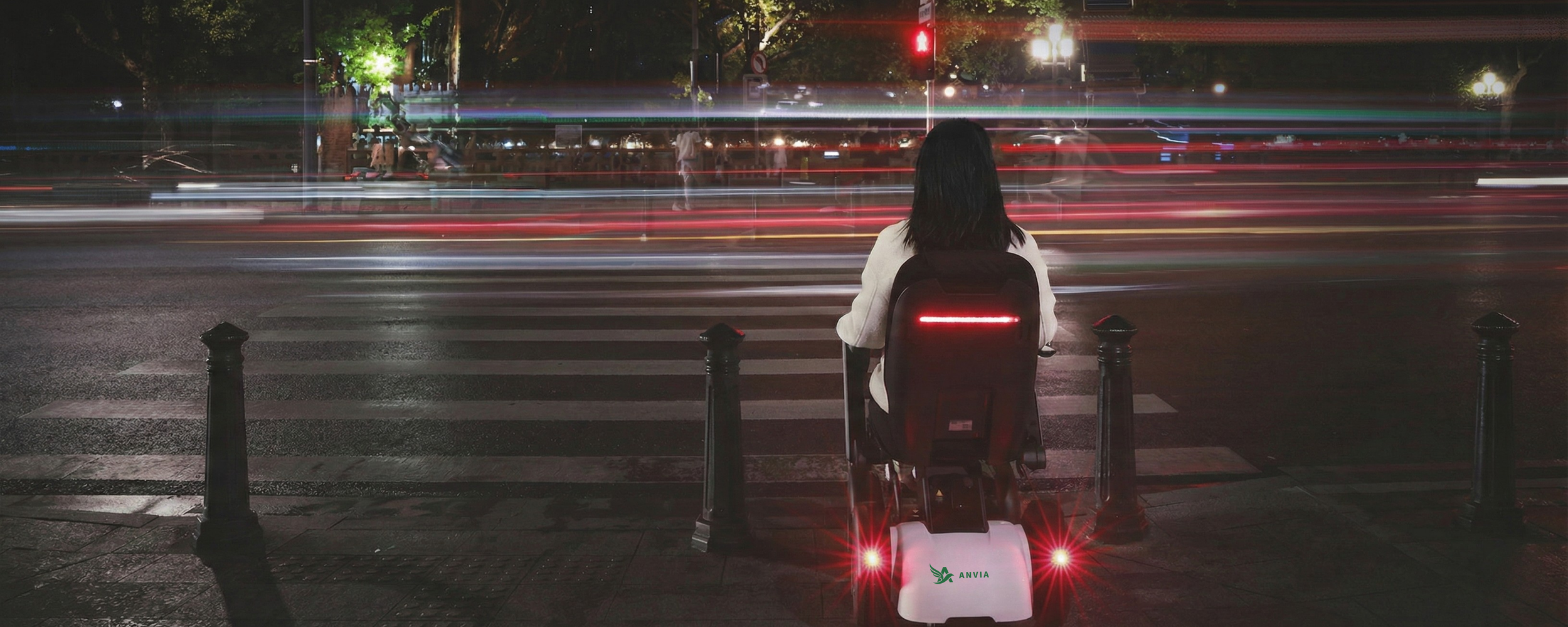 A person sitting on an electric scooter at a crosswalk at night, waiting to cross the street, with light trails from passing vehicles and illuminated traffic signals in the background.