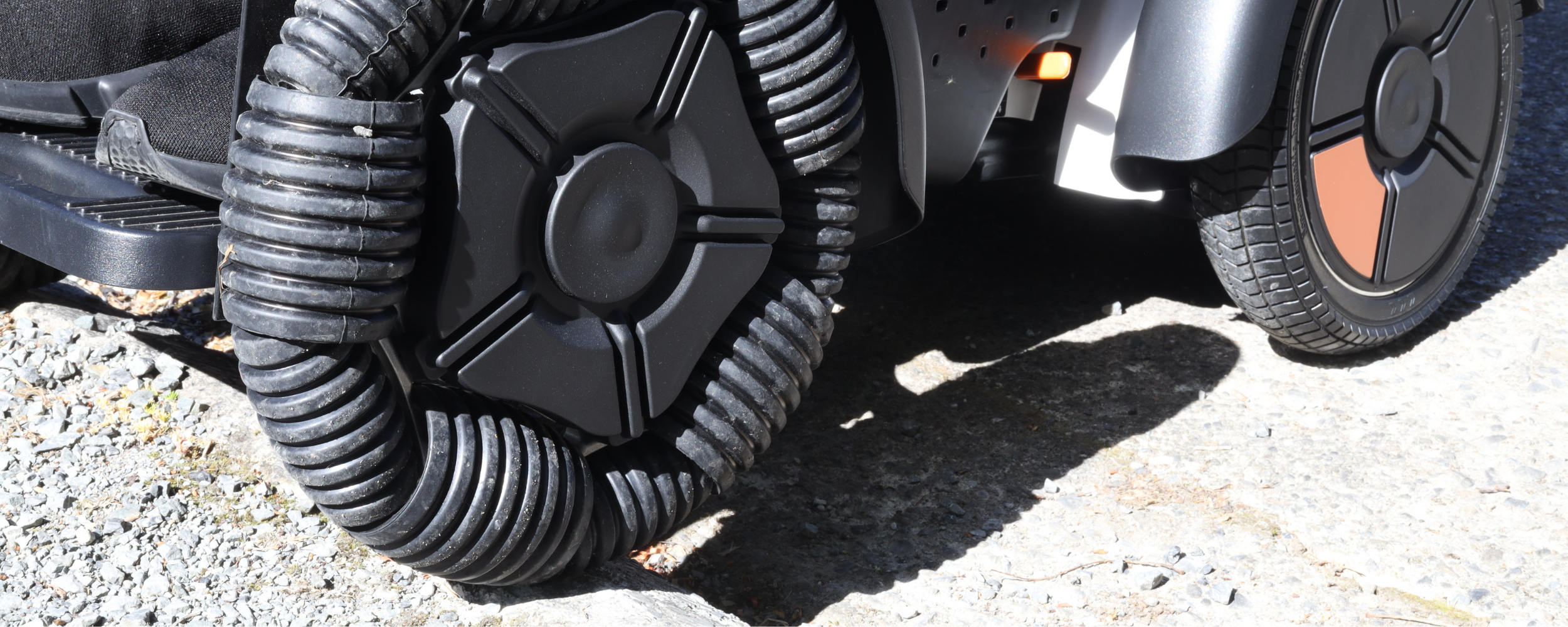 Close-up of a mechanical device on a vehicle with black hoses and a wheel on a gravel surface.