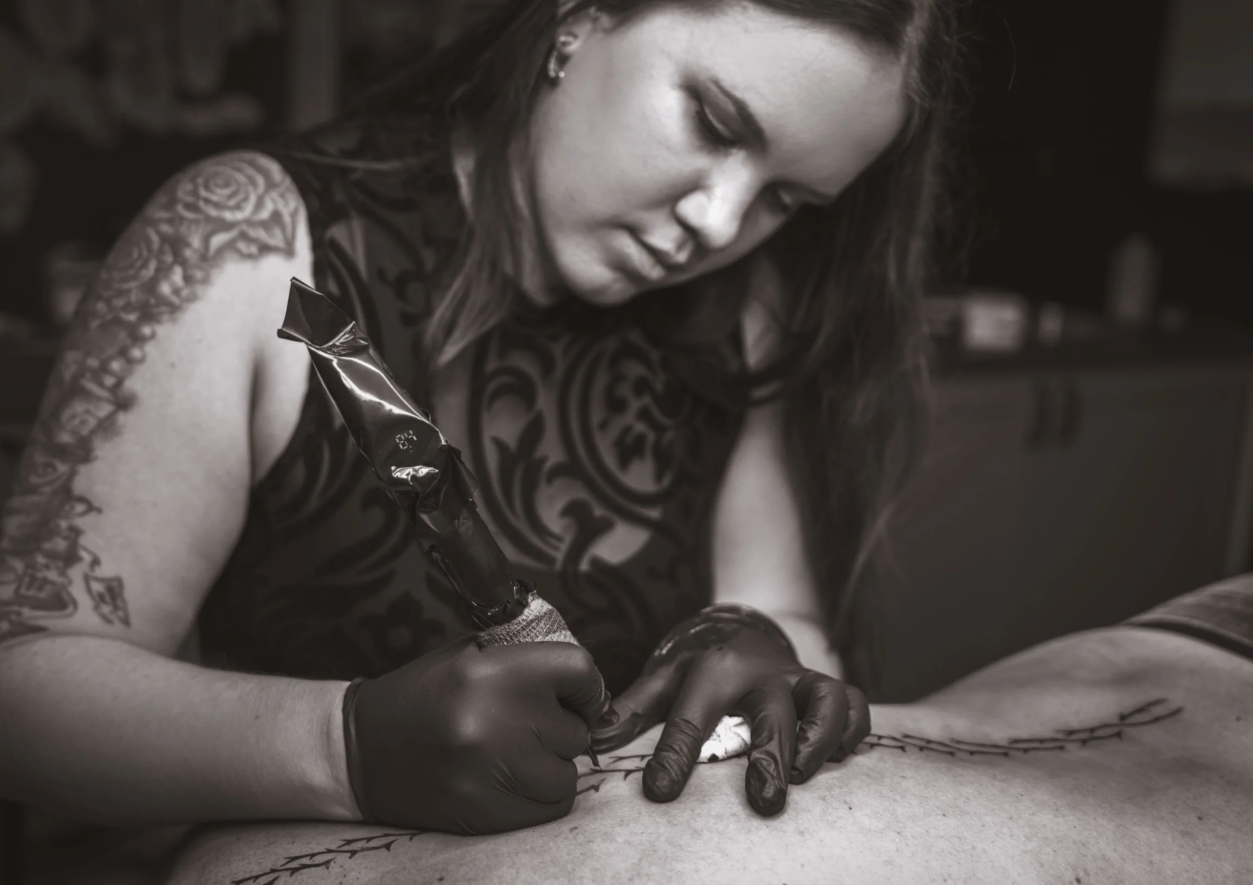Tattoo-Künstlerin arbeitet an einem Tattoo auf einem Kunden, sie trägt schwarze Handschuhe und ein ärmelloses, dunkles Kleid mit großem Muster, in einem Tattoo-Studio.