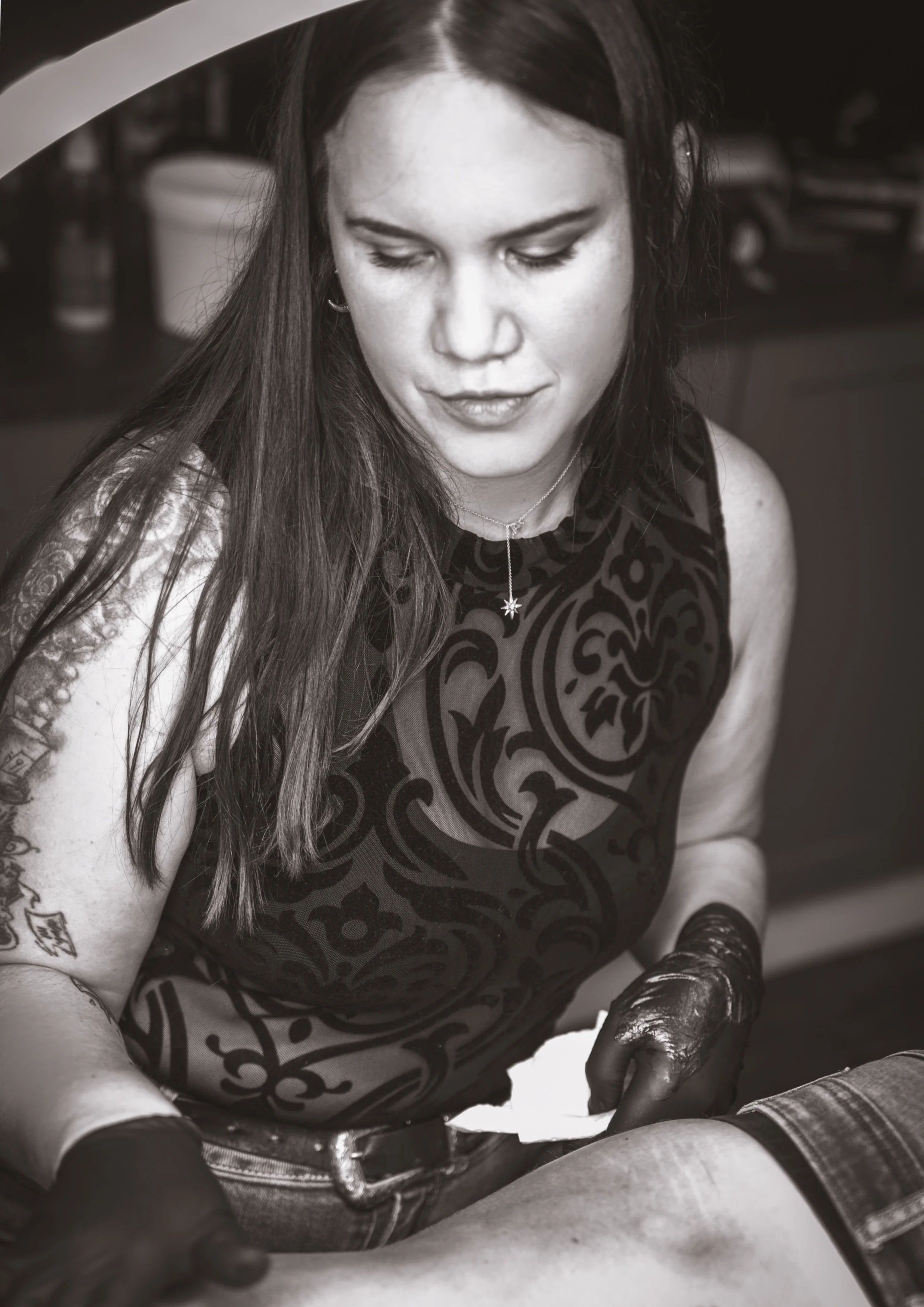 Schwarz-weiß-Foto einer Frau mit Tattoo am Arm, die ein Tattoo auf einer Person macht, während sie Handschuhe trägt.