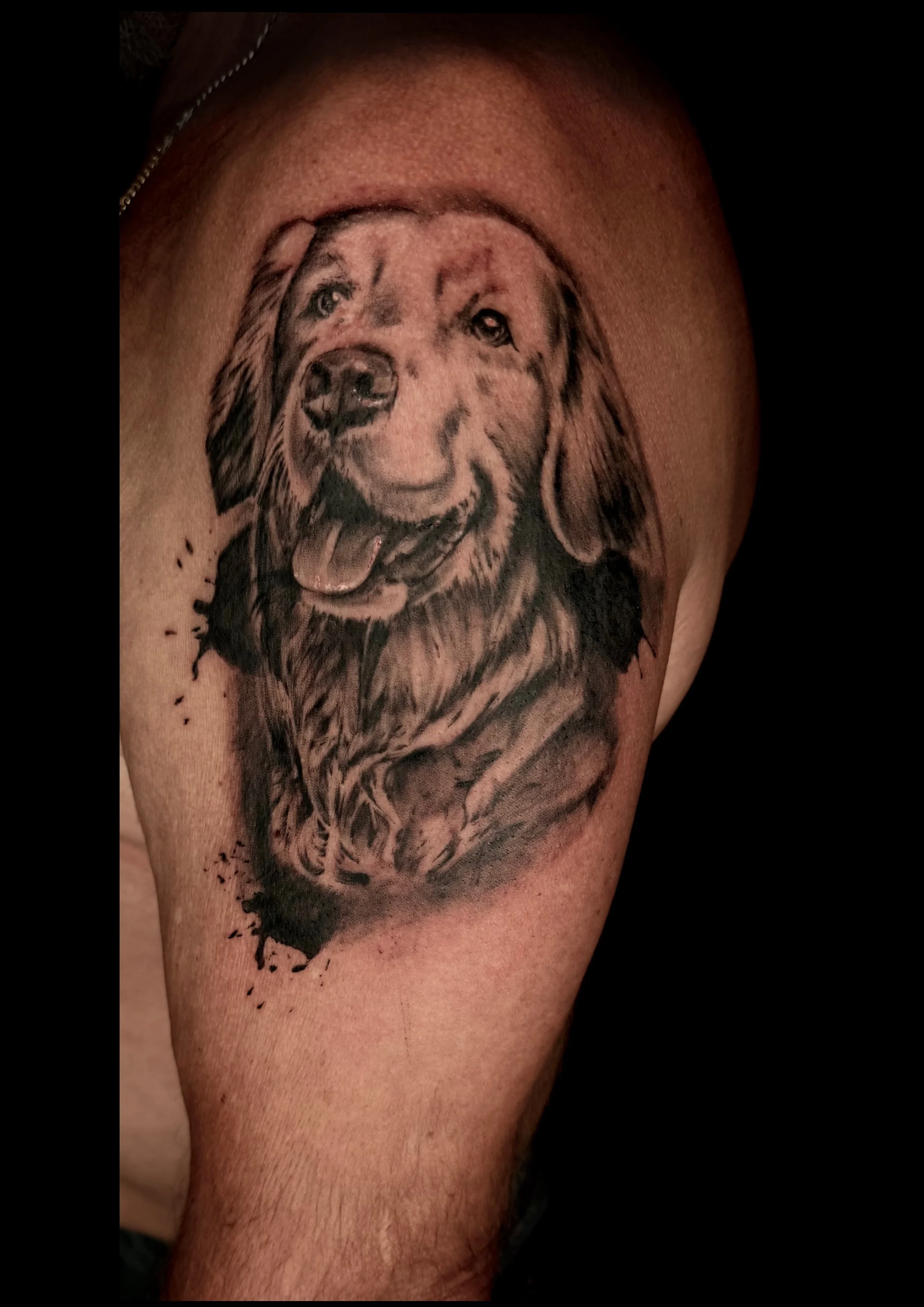 Tattoo eines Hundes auf dem Oberarm, realistisches Design in Schwarzweiß, zeigt einen lächelnden Hund mit offenem Mund und heraushängender Zunge.