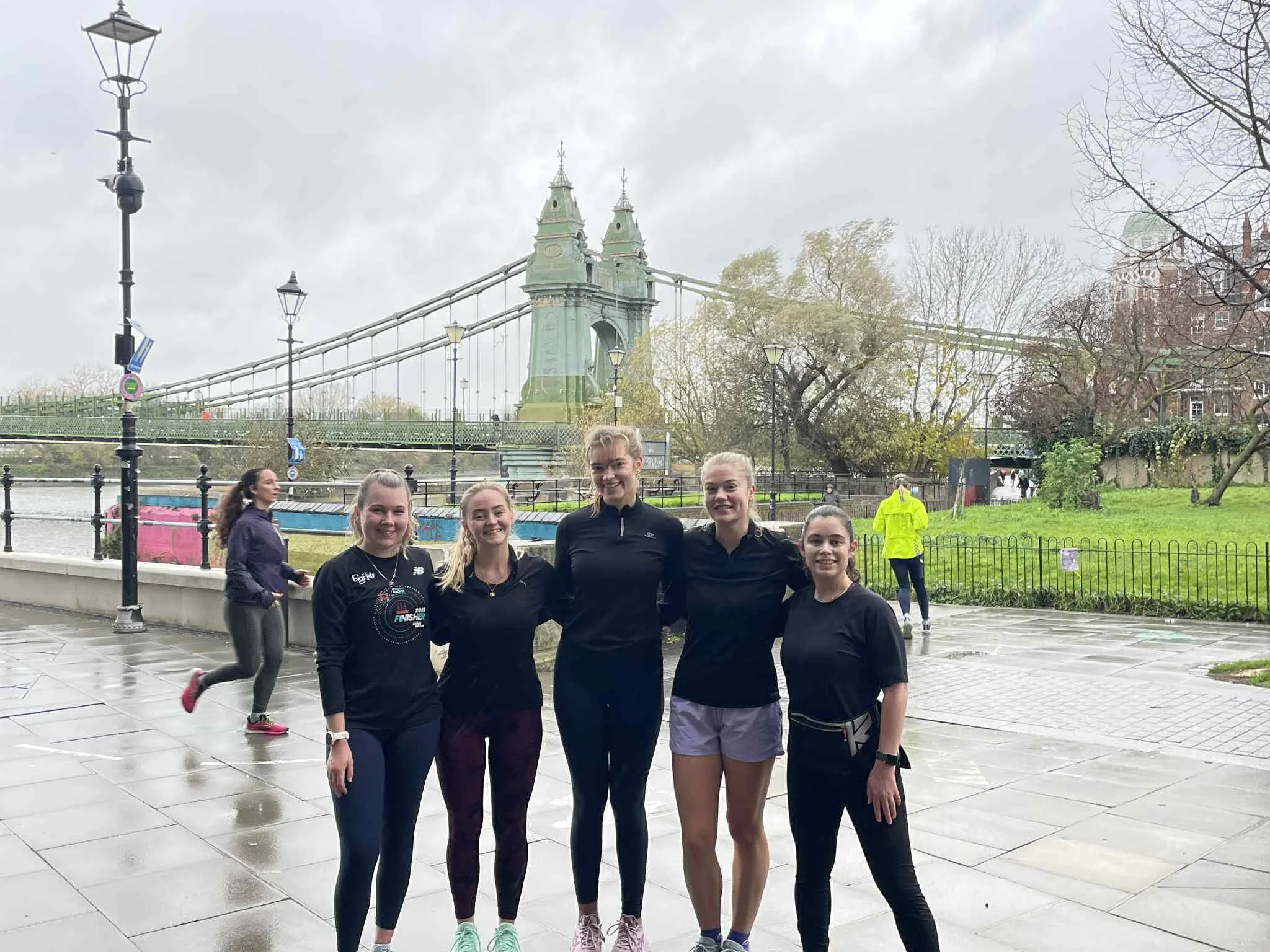 Five women dressed in athletic clothing standing together on a wet sidewalk near a river, with the Tower Bridge in London in the background, overcast sky.