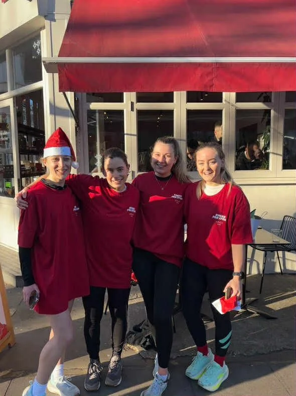 Four women standing outside a building, smiling, wearing red shirts and one woman in a Santa hat. They appear to be participating in a festive event.